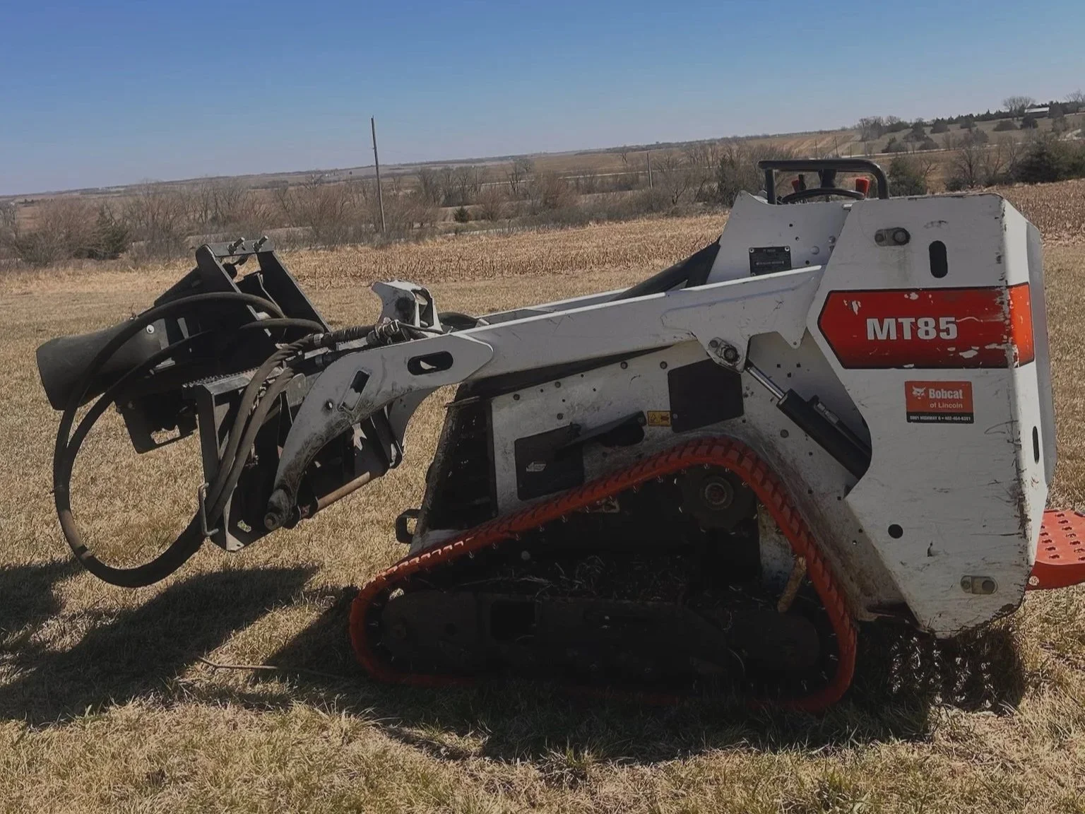 A white and orange Bobcat MT85 compact track loader with a bucket attachment, sitting on a grassy field with a rural landscape and power lines in the background.