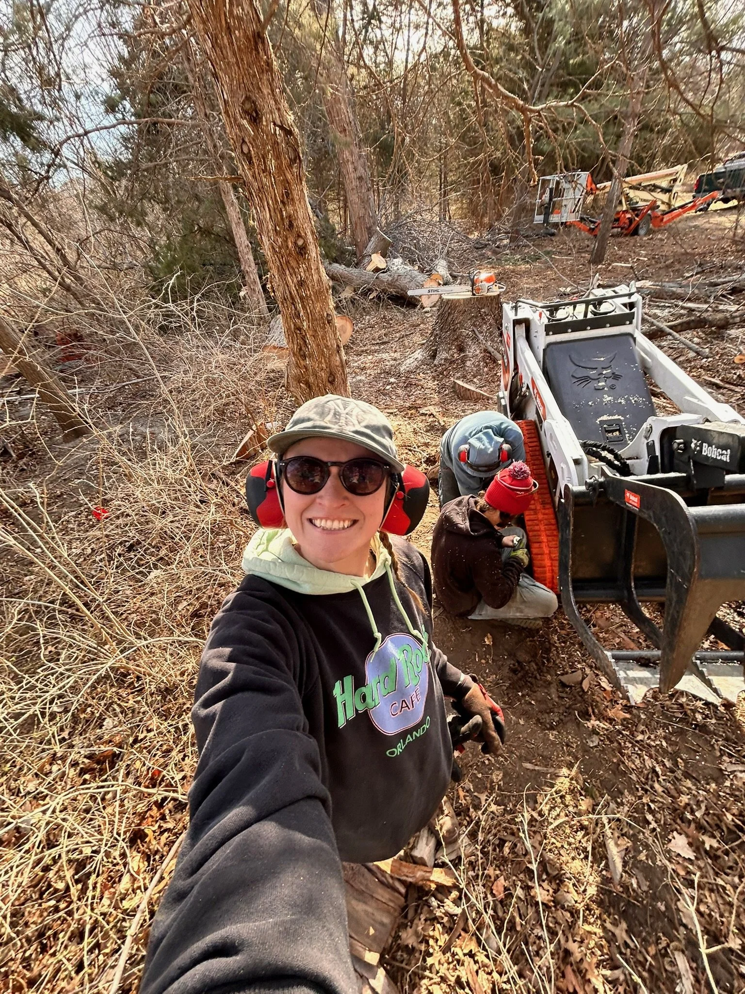 A woman wearing safety glasses, a Hard Rock Cafe Orlando hoodie, and red earmuffs taking a selfie in a forested area with trees, fallen branches, and two people working with chainsaws in the background.