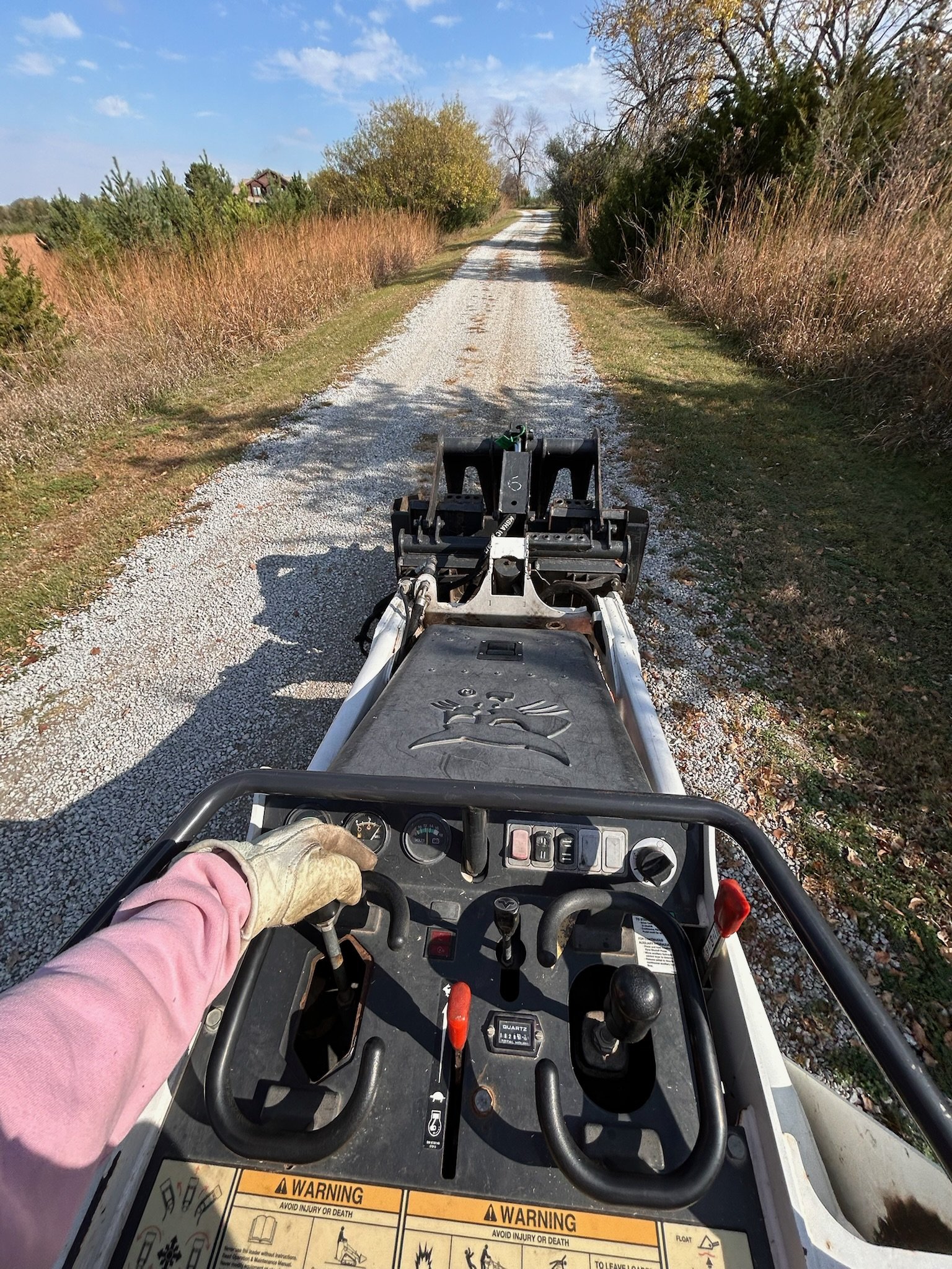 First person view from a skid steer loader (or similar construction vehicle) operator's seat, holding the steering wheel, overlooking a gravel dirt road with trees on both sides, on a clear sunny day.