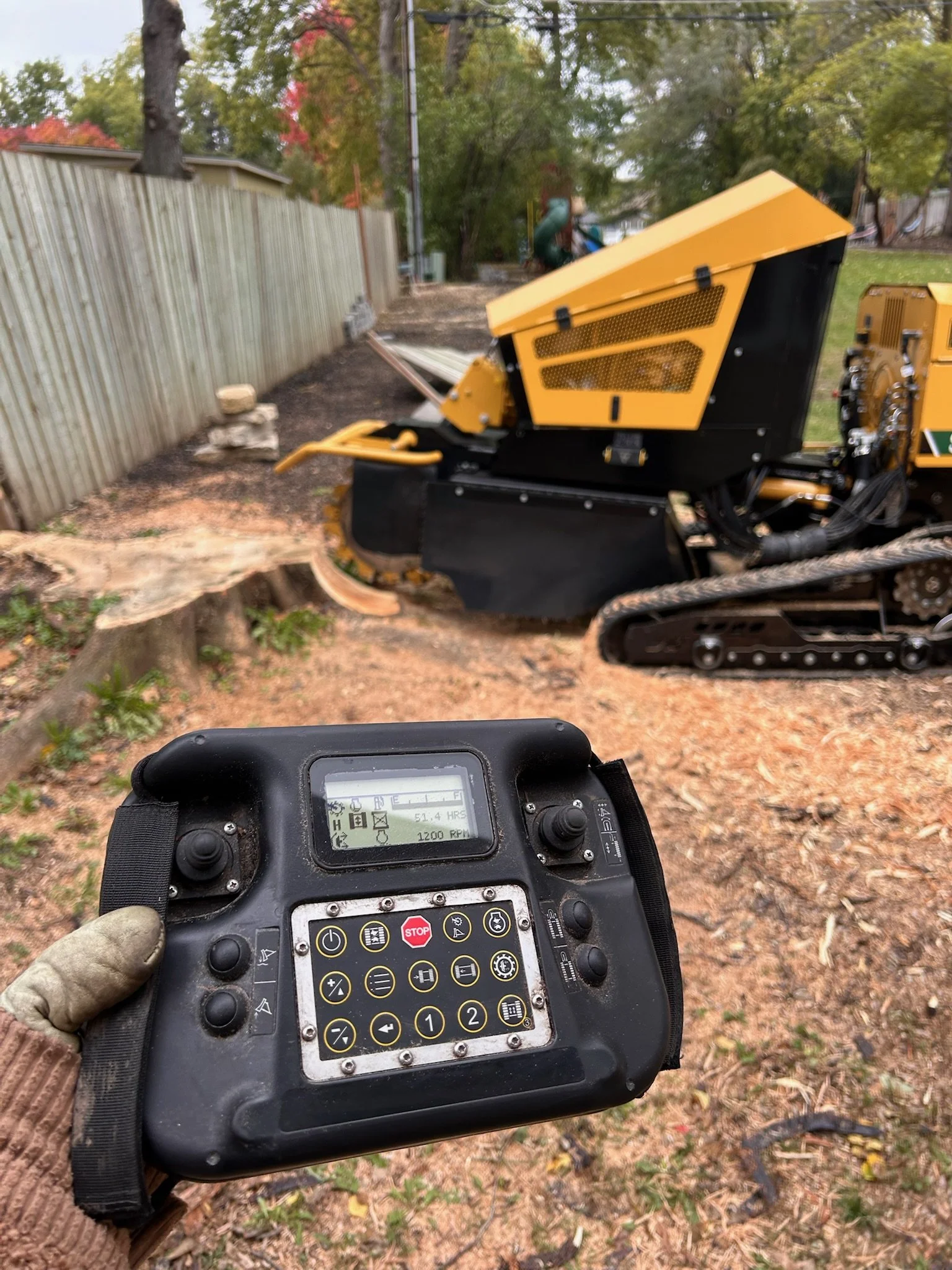 A person holding a remote control device while overseeing a yellow and black tracked wood cutting machine at a yard with a wooden fence and trees in the background.