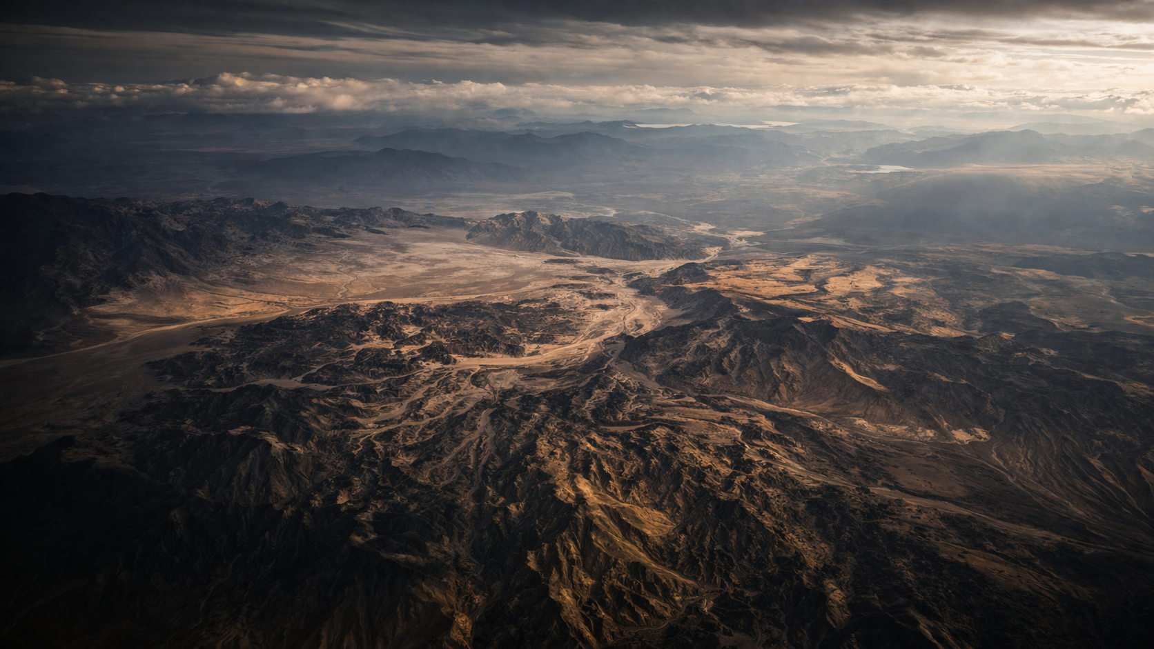 Aerial view of a rugged, mountainous desert landscape with dry riverbeds, mountains, and plains under a cloudy sky.