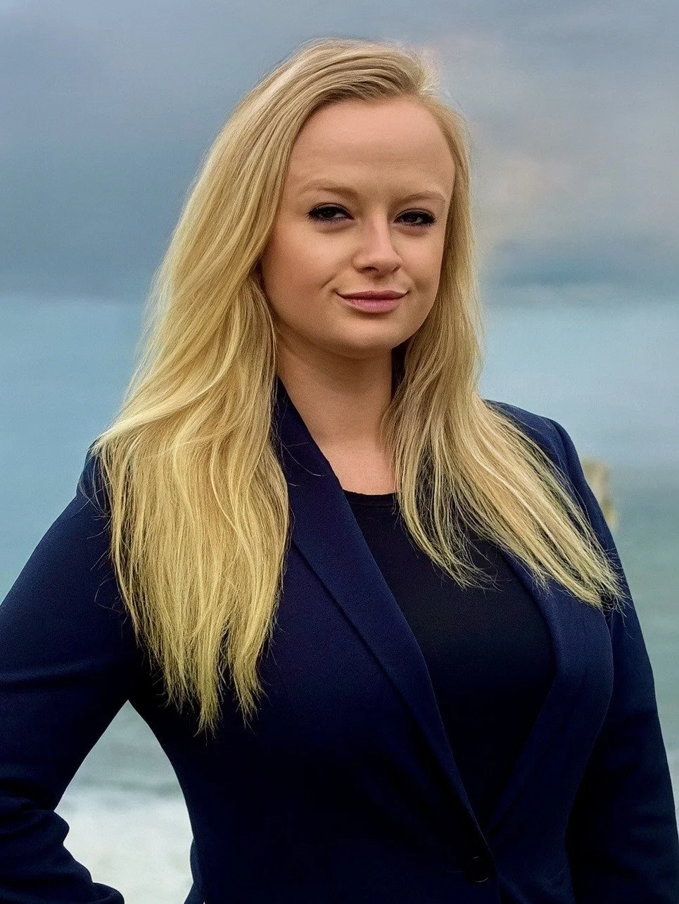 A woman with long blonde hair wearing a navy blazer stands outdoors near the ocean with cloudy skies in the background.