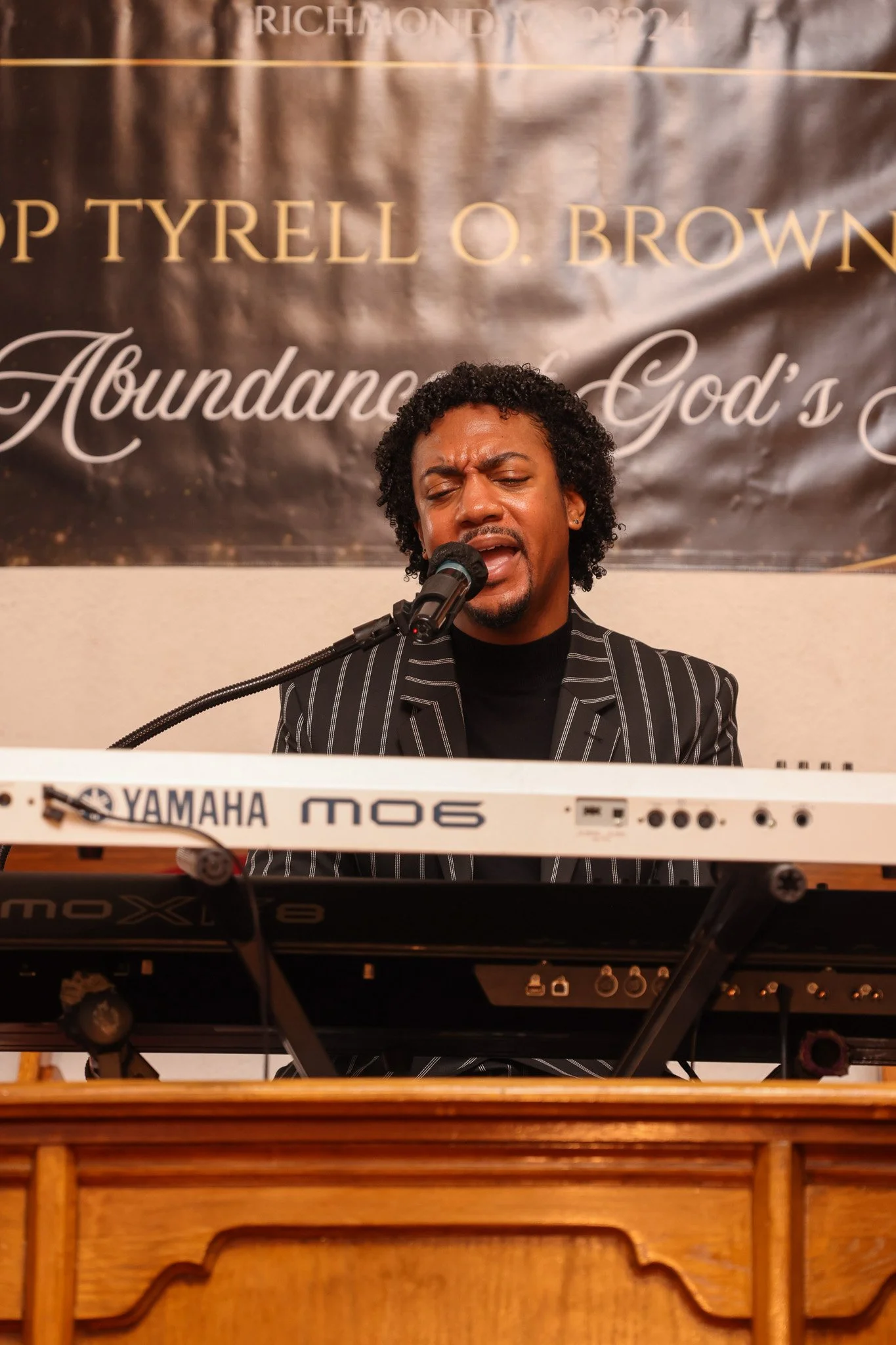 A man with curly black hair singing into a microphone while playing a Yamaha MOX6 keyboard. He is wearing a black and white striped blazer. In the background, there is a banner with text including "Richmond 2021" and opportunity for a personal or church event.