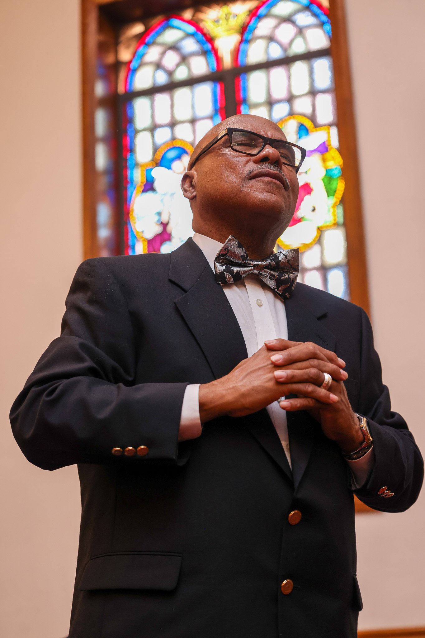 A bald man wearing glasses, a black tuxedo, and a patterned bow tie standing in front of a stained glass window, with his hands clasped together.