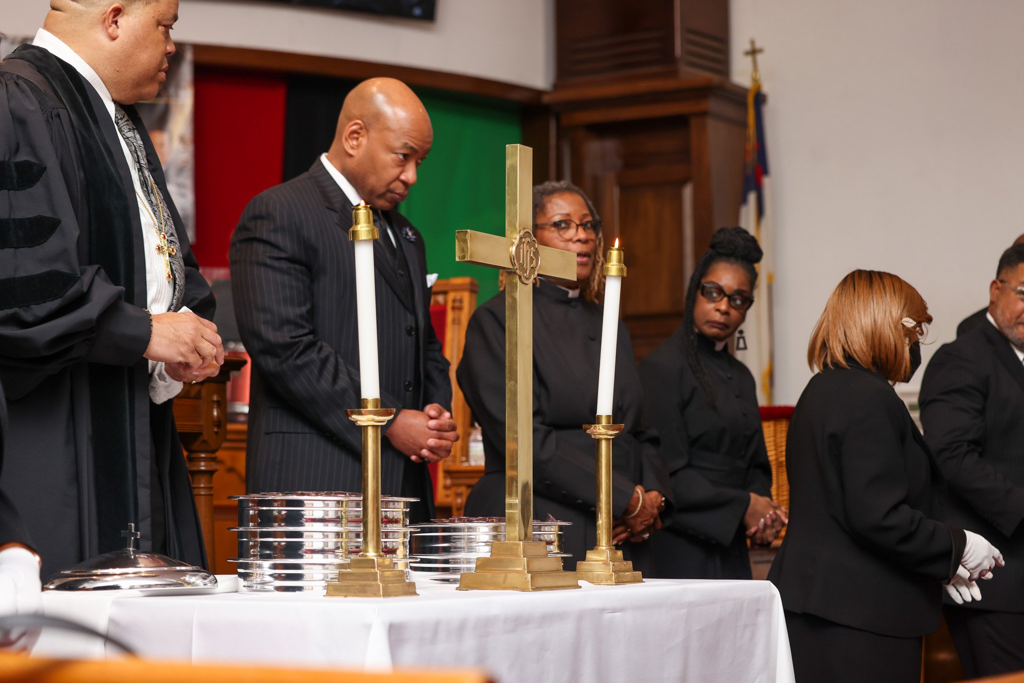 A group of people, mostly dressed in black robes and suits, are standing in a row during a religious ceremony in a church. In the foreground, there are tall, gold-colored candlesticks with lit candles on a white table. The background shows a wooden cross and flags.