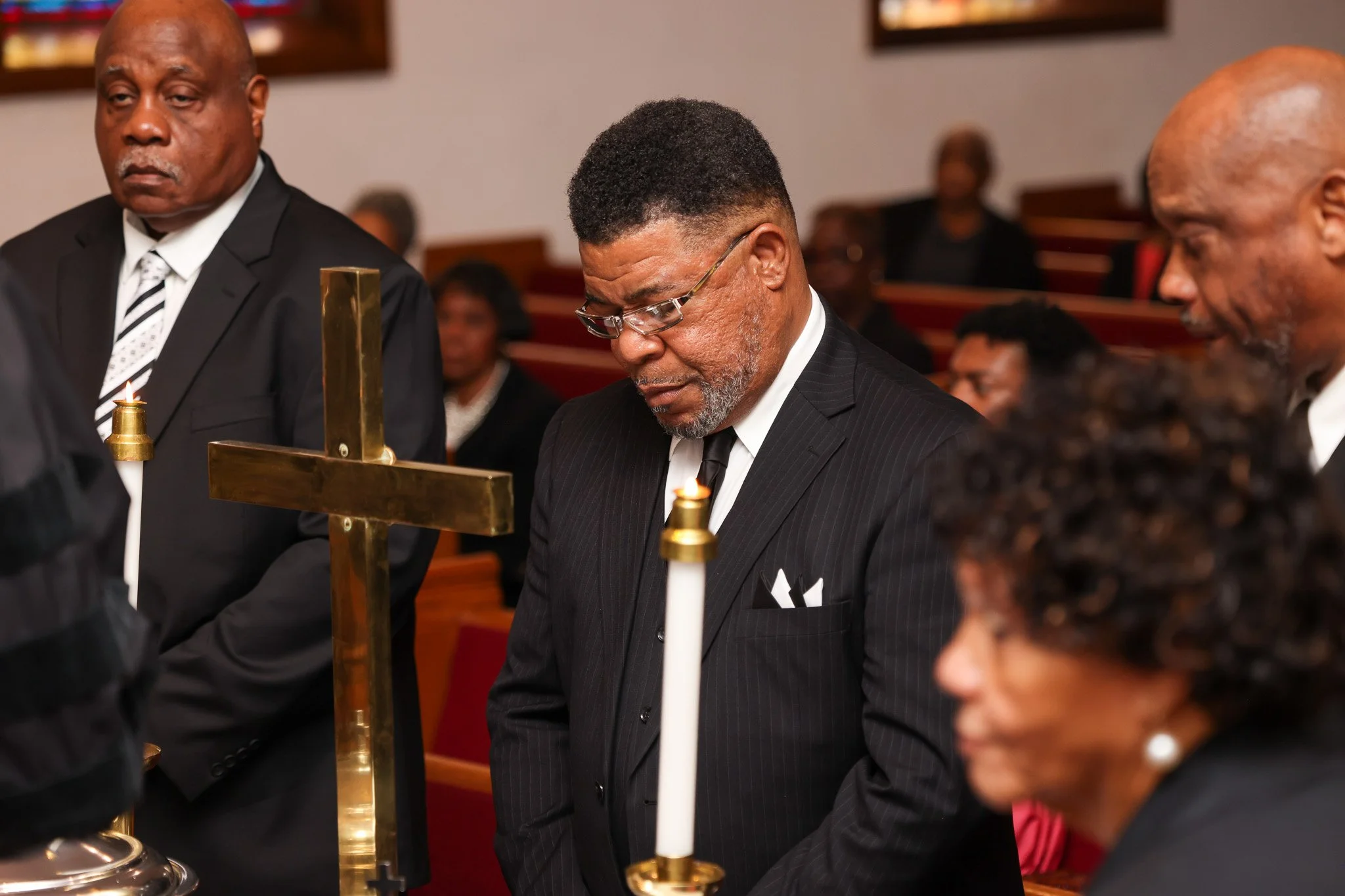 Three men wearing black suits and white shirts are gathered around a brass candle holder during a religious or memorial service in a church. One man is holding a lit candle, while a cross is visible on the candle holder. The background shows other attendees and wooden pews.