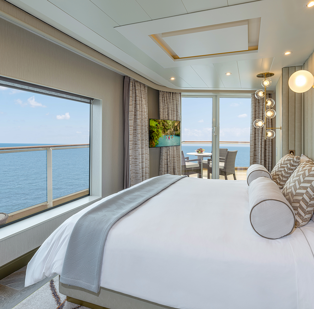 Modern bedroom with large window and sliding door overlooking the ocean, with a balcony table and chairs, and a flat-screen TV.