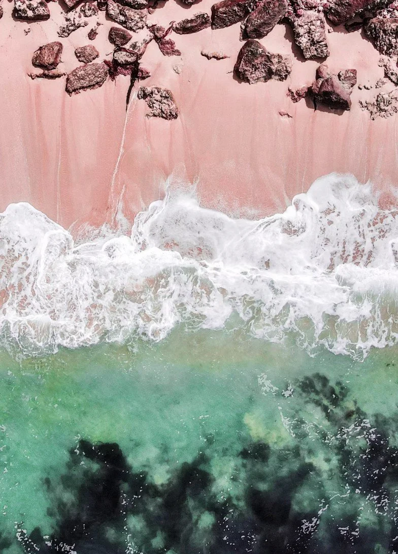 Overhead view of a pink beach with rocks at the top and waves crashing onto the shore below, turquoise water with dark patches at the bottom.