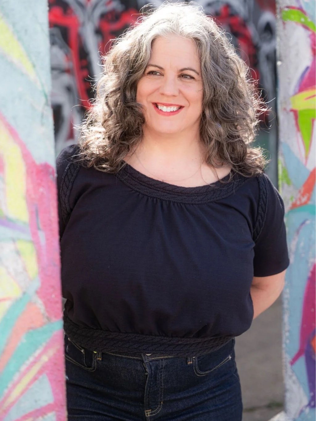 A woman with curly, two-toned gray and brown hair smiling outdoors, standing between two graffiti-covered walls.