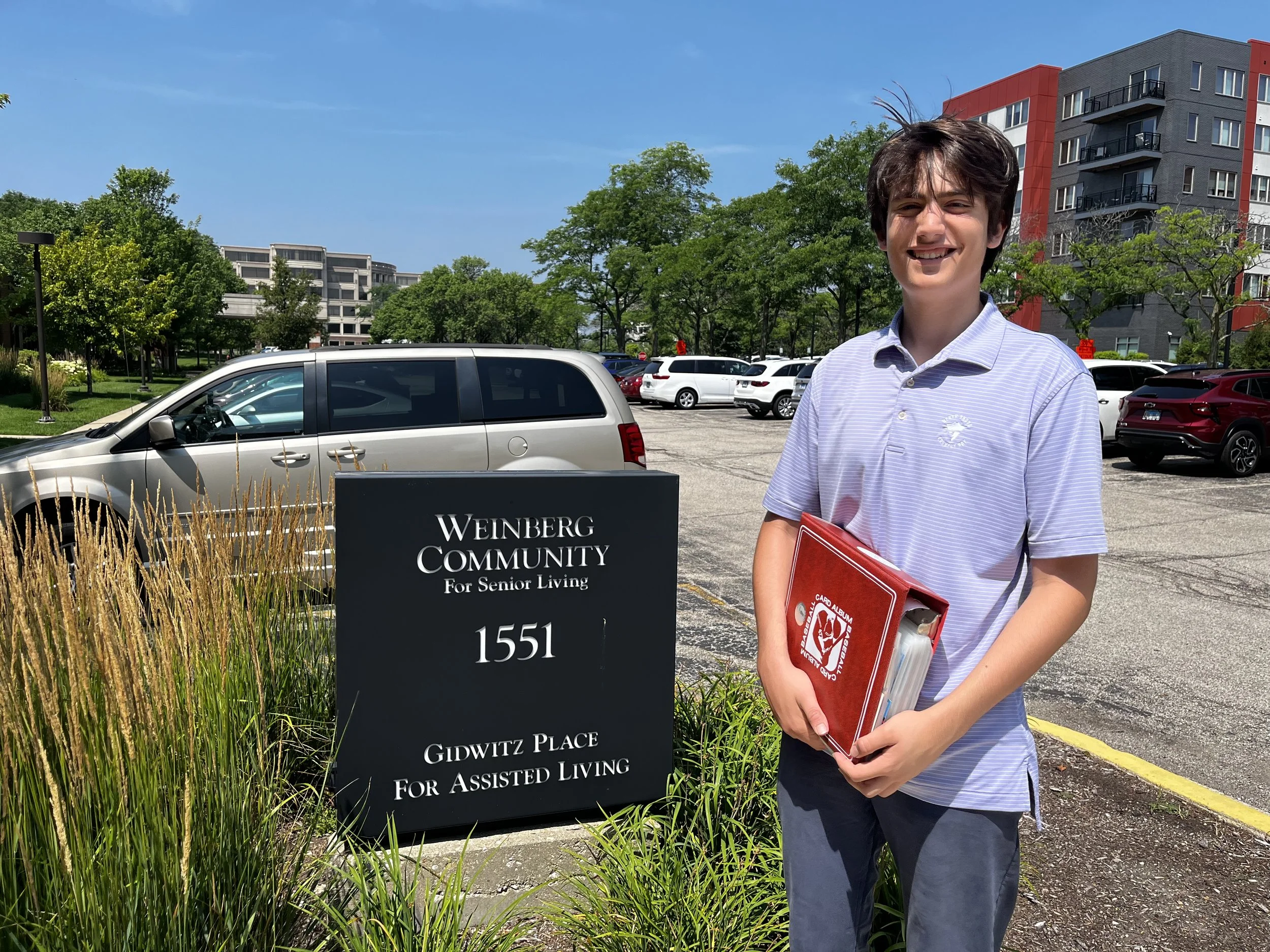 A young man standing outdoors in a parking lot, holding a red folder and smiling. Behind him is a sign for Weinberg Community for Senior Living at 1551 Gidwitz Place, with trees, parked cars, and apartment buildings in the background.
