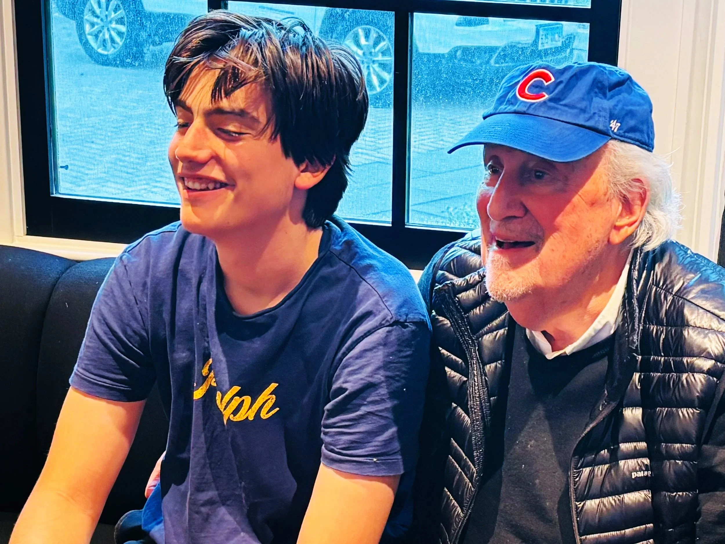 Two men sitting on a black couch near a window, smiling and enjoying each other's company. The younger man has dark hair and is wearing a blue t-shirt, while the older man has white hair, a beard, and is wearing a blue baseball cap with a red 'C', a black puffer jacket, and a gray sweater.