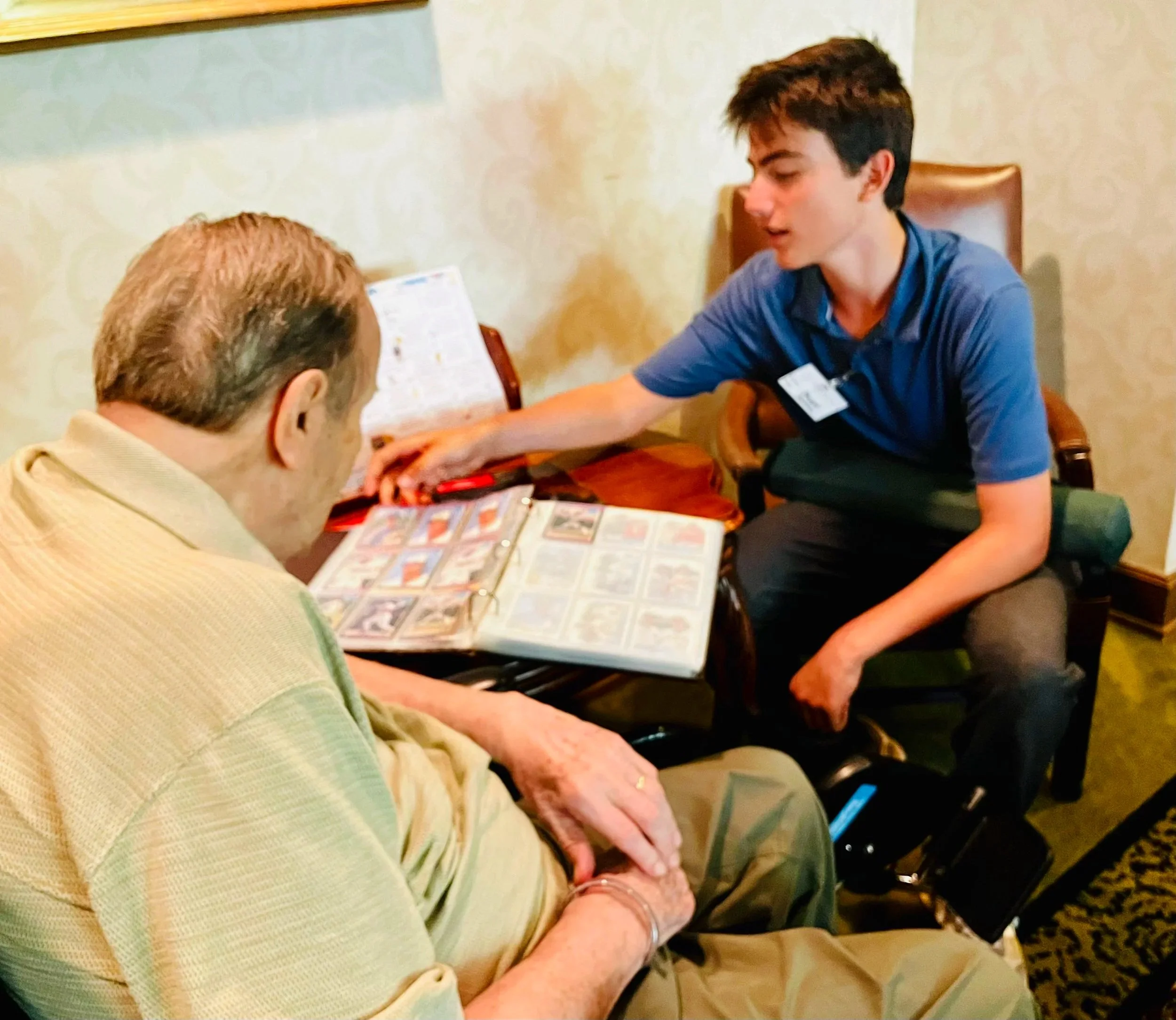 A young man in a blue polo shirt showing photo albums to an elderly woman in a beige shirt in a cozy room with patterned wallpaper.