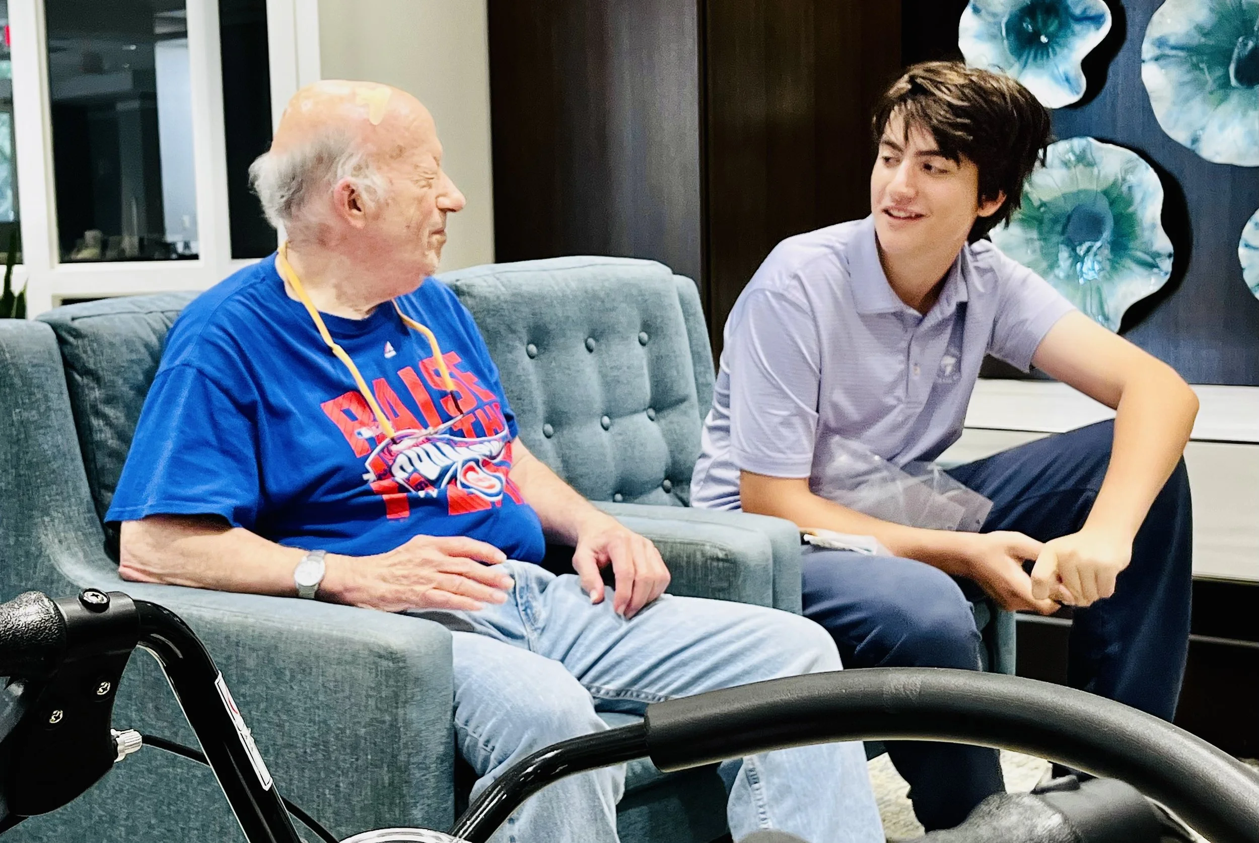 An elderly man with a walker seated on a teal sofa, talking with a young man in a light purple shirt, in a living room with decorative wall art.