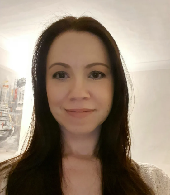 A woman with long dark hair smiling in a well-lit room with a painting or photograph on the wall behind her.