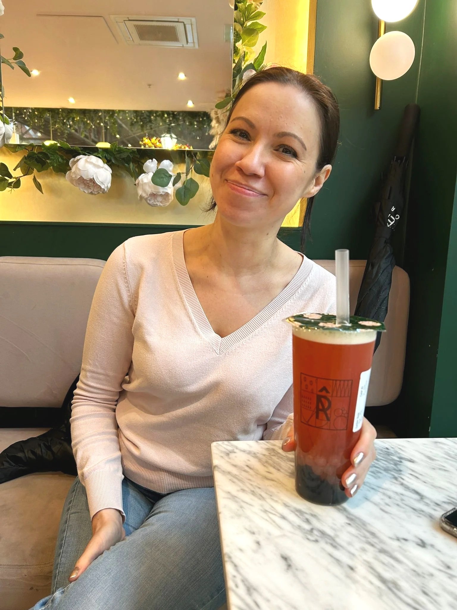 Woman sitting at a marble table in a restaurant or cafe, holding a large beverage in a clear cup with a straw, decorated with a label and lid, smiling.