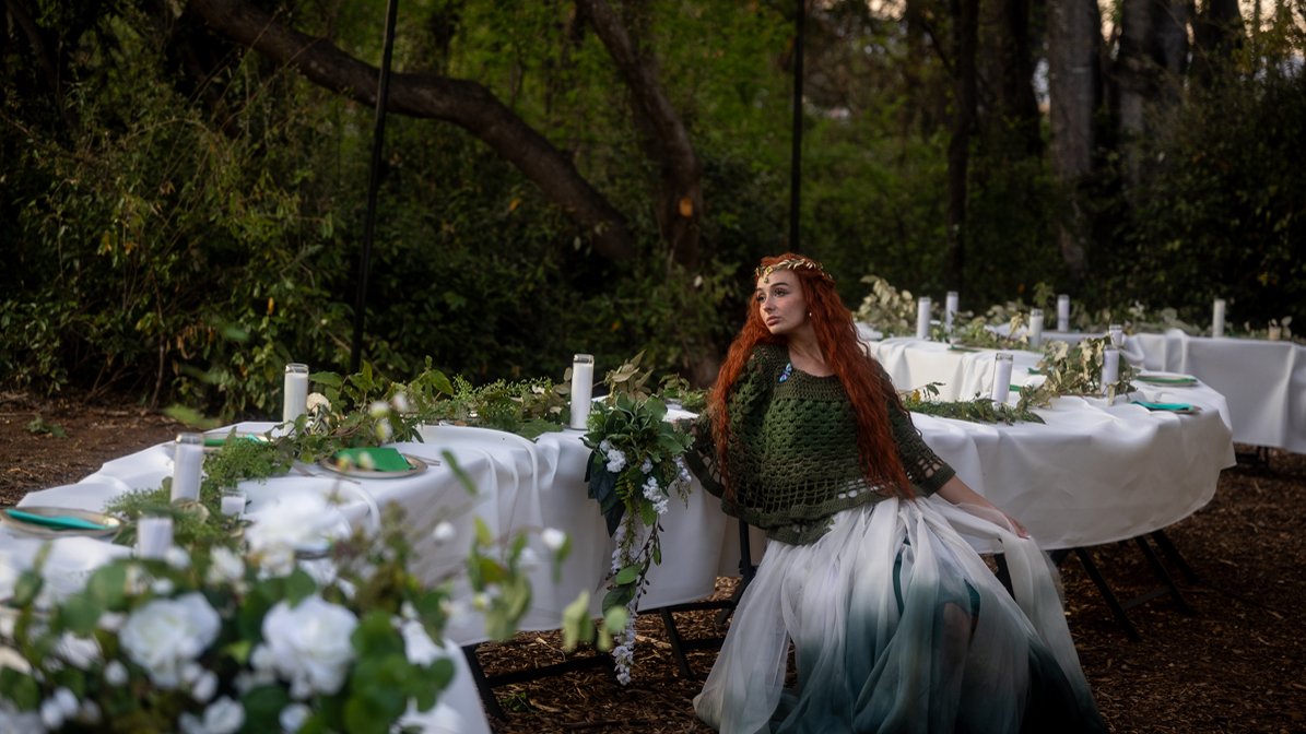 A woman with red hair sitting at a long outdoor dining table decorated with white tablecloths, candles, greenery, and white flowers, surrounded by trees and nature.