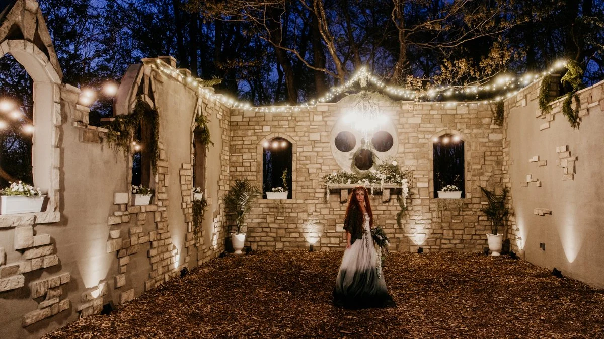 A woman stands in a haunted-themed outdoor setting decorated with string lights, a large skull mural above a brick fireplace, and potted plants, during twilight or early night.