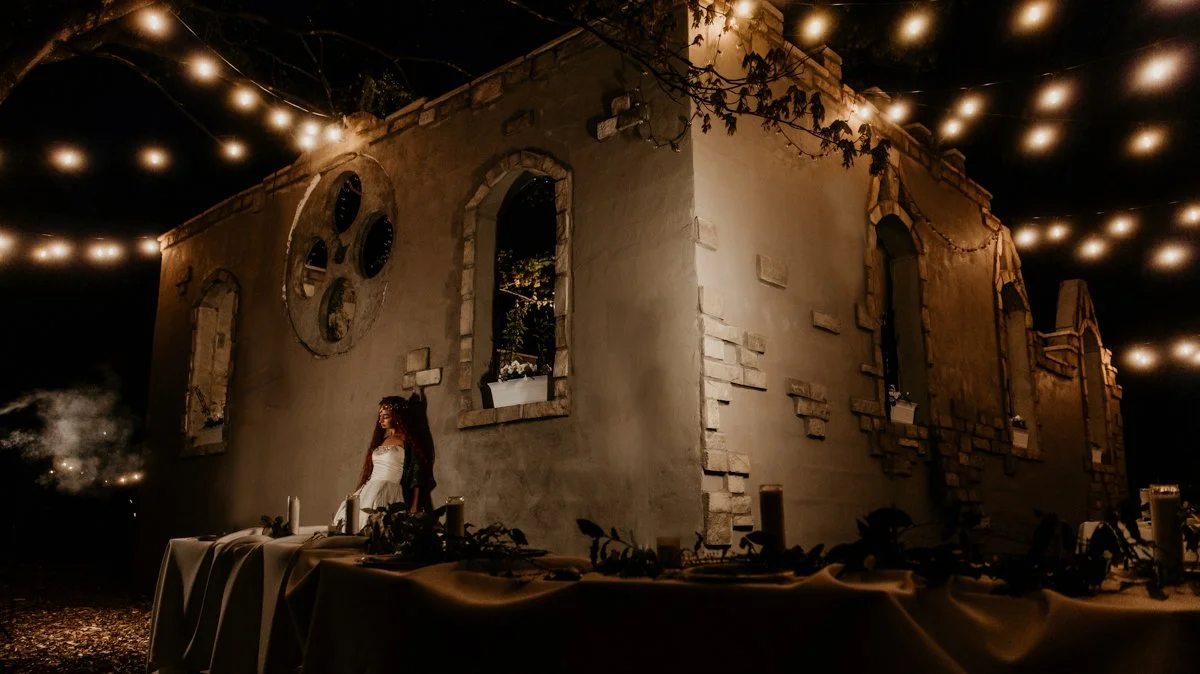 Nighttime scene with a woman in a white dress standing beside a table set with candles and decorations in front of an old stone building with tall, arched windows. String lights are hanging above, illuminating the scene.