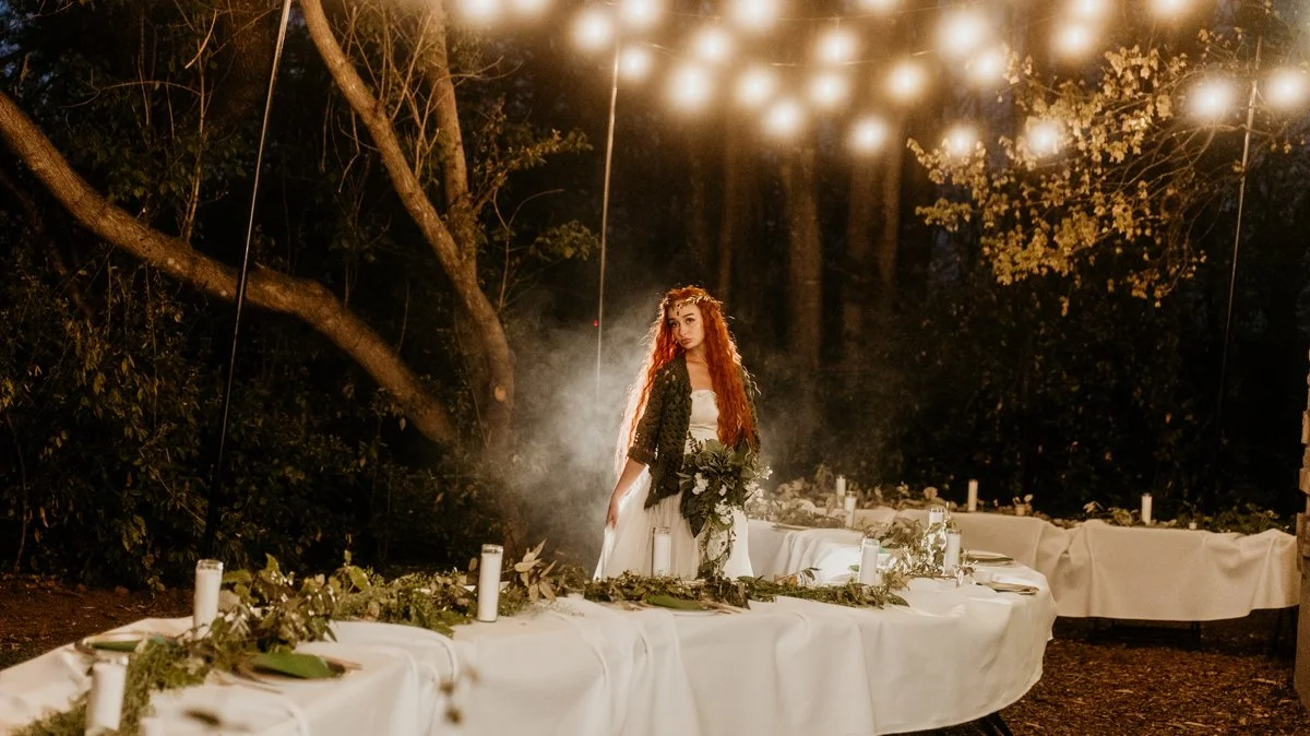 A woman with long red hair in a forest setting, standing behind a table decorated with greenery and candles, illuminated by string lights overhead at night.
