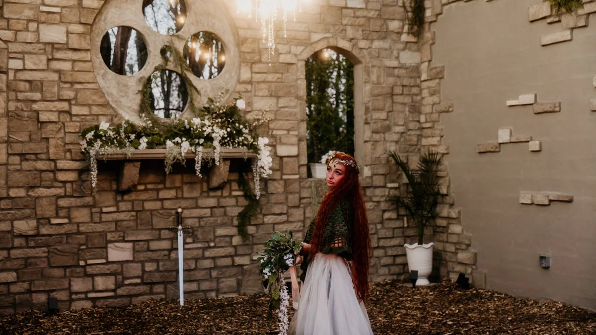 A woman with long red hair in a white dress and a green sweater, holding a bouquet, stands in front of a stone wall decorated with white flowers and a large circular art installation.