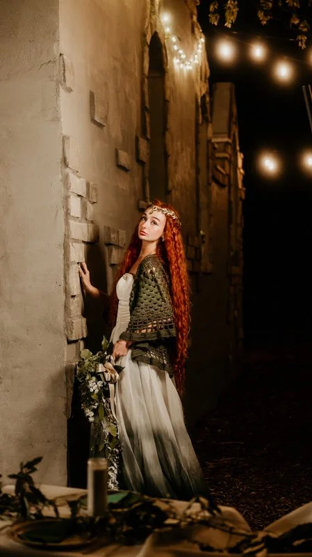 A young woman with long, curly red hair standing outdoors at night, beside a stone wall decorated with string lights, holding a bouquet of flowers, wearing a flowing white dress and a dark green shawl.