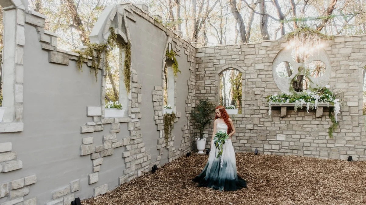 A woman in a strapless white dress with a dark ombré effect at the bottom holds a bouquet of greenery and white flowers inside a rustic stone-walled outdoor space with leaf-covered ground and some plants, with sunlight filtering through the trees.