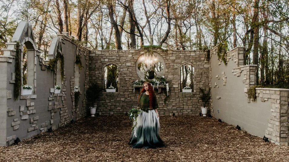 A woman in a long green and white gradient dress holding a bouquet of flowers stands inside a ruined stone building with arched windows, decorated with flowers, with trees in the background.