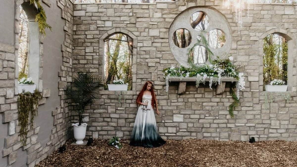 A woman with long red hair wearing a white and black ombre dress stands in front of a stone wall decorated with white flowers and greenery. There are three small windows or openings in the wall, with potted plants on the ledges, and a large round dec