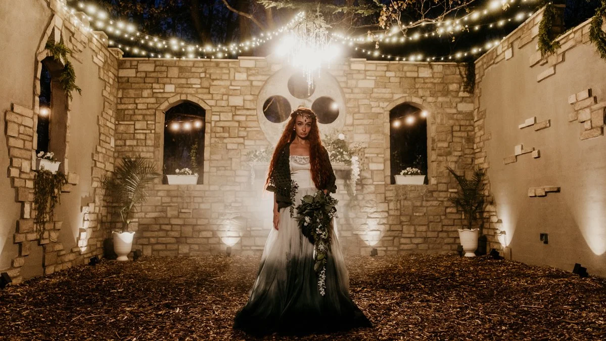 A woman in a white and black dress holding a bouquet stands in an outdoor decorated area with string lights, potted plants, and a stone wall.