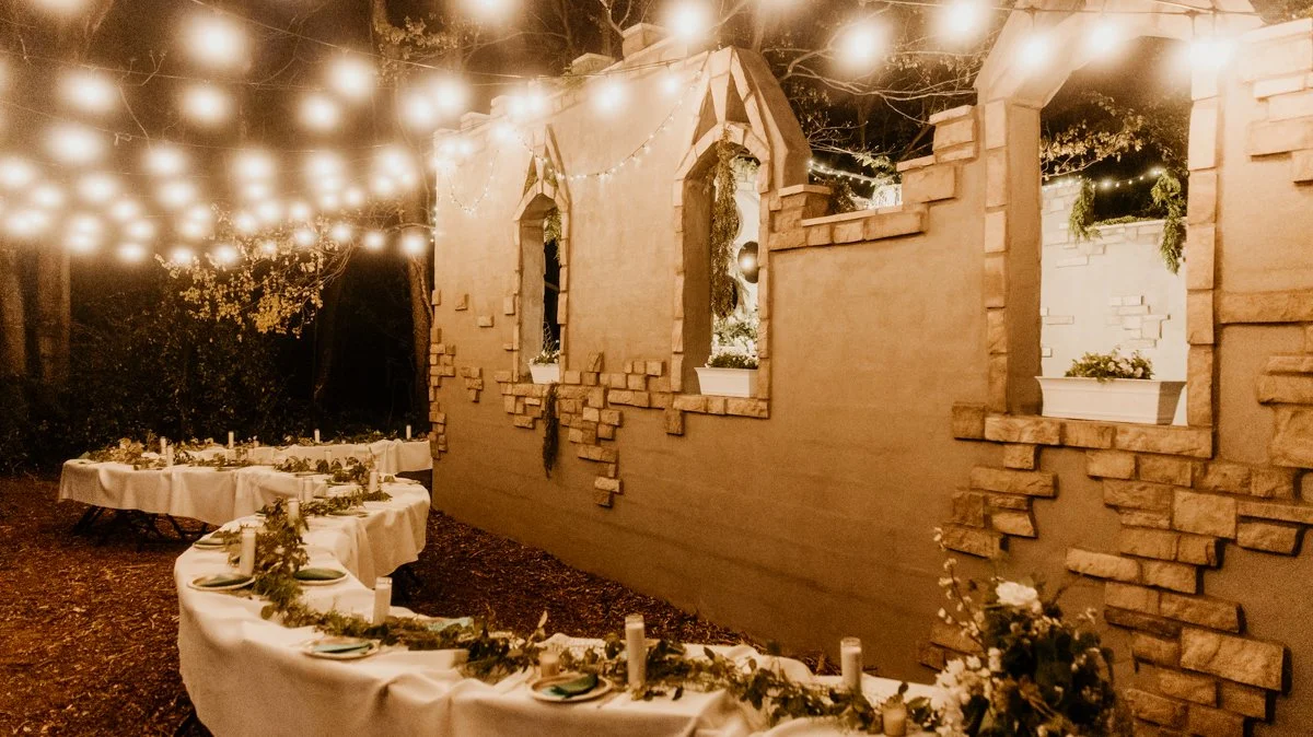 Outdoor evening gathering with string lights, decorated tables, and window boxes with plants against a brick wall.