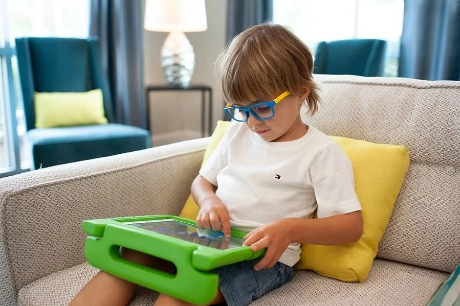 A young boy with glasses sitting on a couch, using an AAC communication app on a green tablet during in-home speech therapy for kids in Pewaukee, WI.