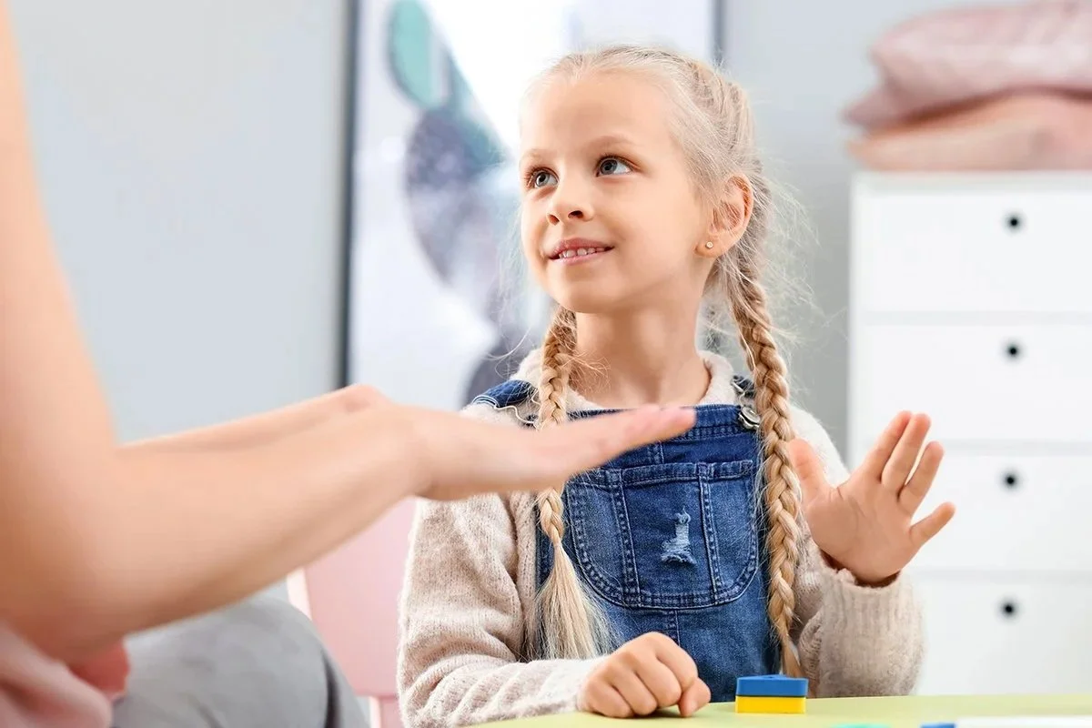 Young girl with blonde braids and denim overalls smiling during in-home speech therapy for kids in Waukesha County, WI.