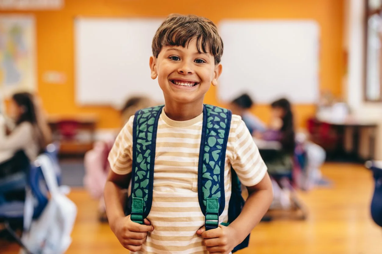 A smiling young boy with a backpack in a classroom with other children in the background, with improved social skills because of speech therapy provided by a school contract speech therapist in Wisconsin.