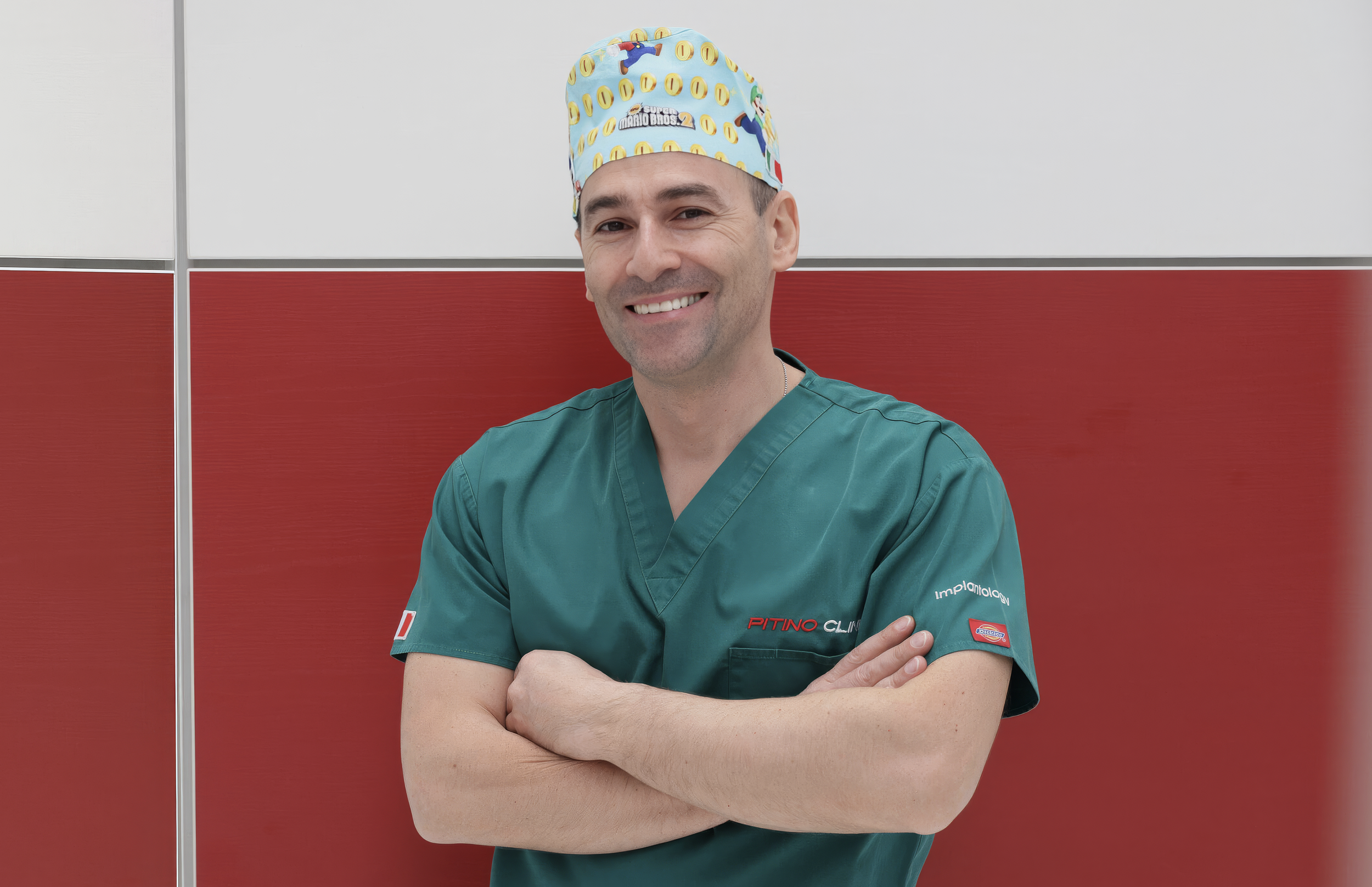 Un medico sorridente con camice verde e cappello con personaggi di Super Mario Bros.2, in piedi davanti a uno sfondo di pannelli grigi e rossi.