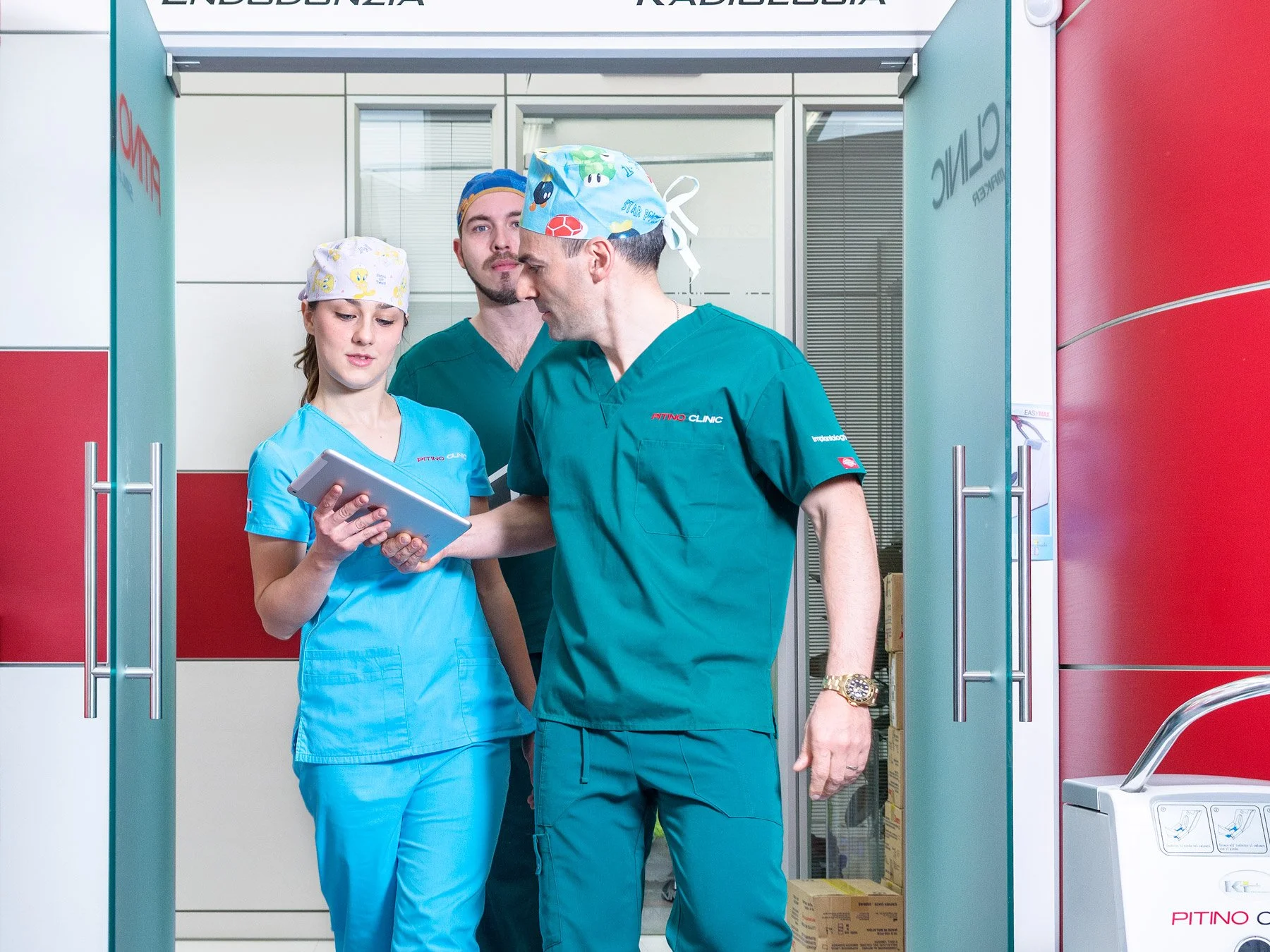 Tre medici e un'infermiera in corsia d'ospedale, indossano camici e cappelli chirurgici, e sembrano discutere o consultarsi su dispositivi digitali.