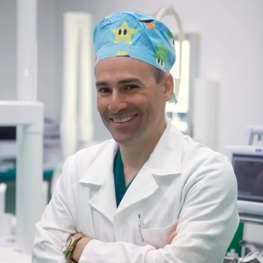 Un medico sorridente indossa un camice bianco e un cappello chirurgico con disegni di stelle e dinosauri, in un ospedale.