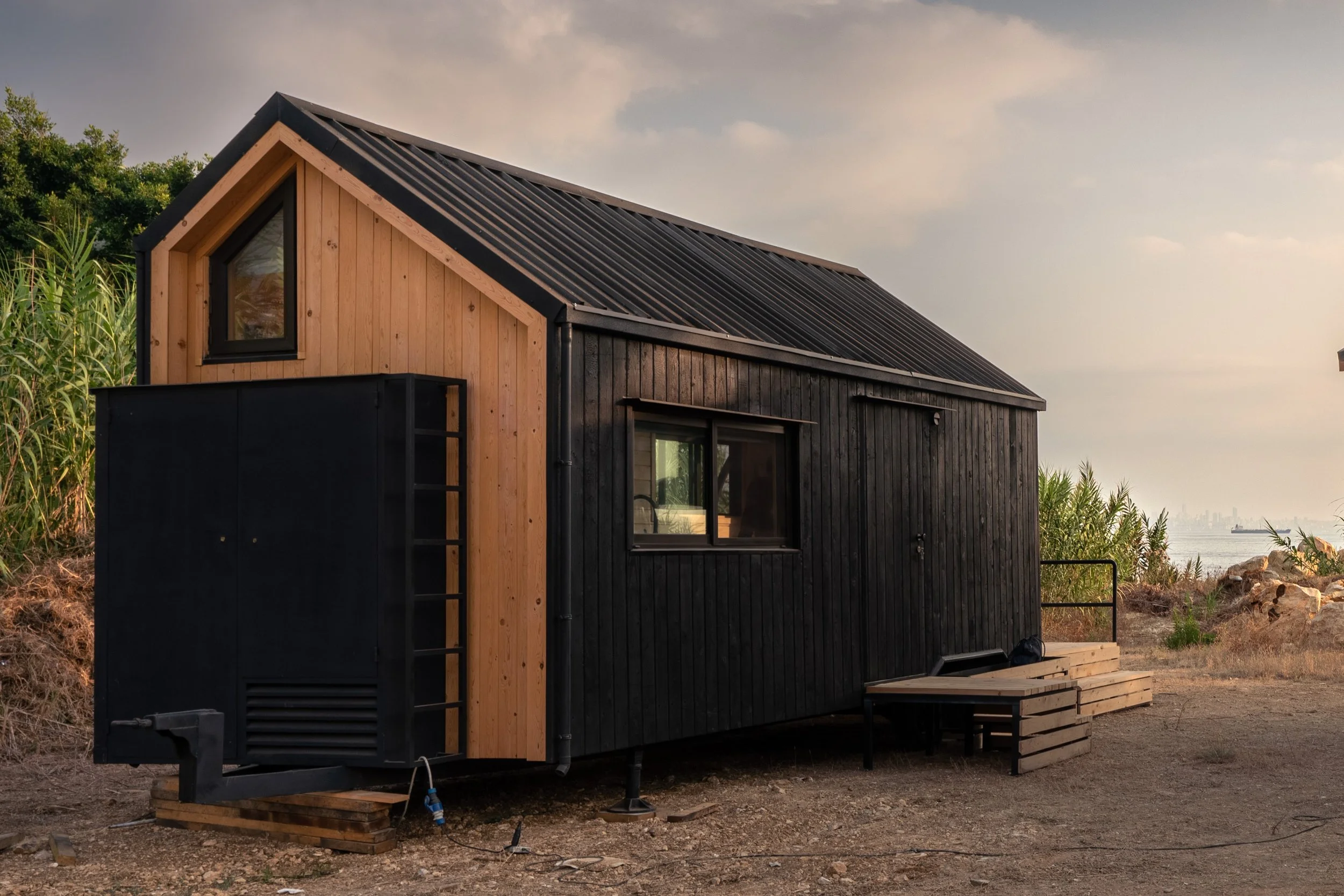 A modern tiny house with black and natural wood siding, a sloped metal roof, and large windows, built on a raised platform in a natural setting near water.