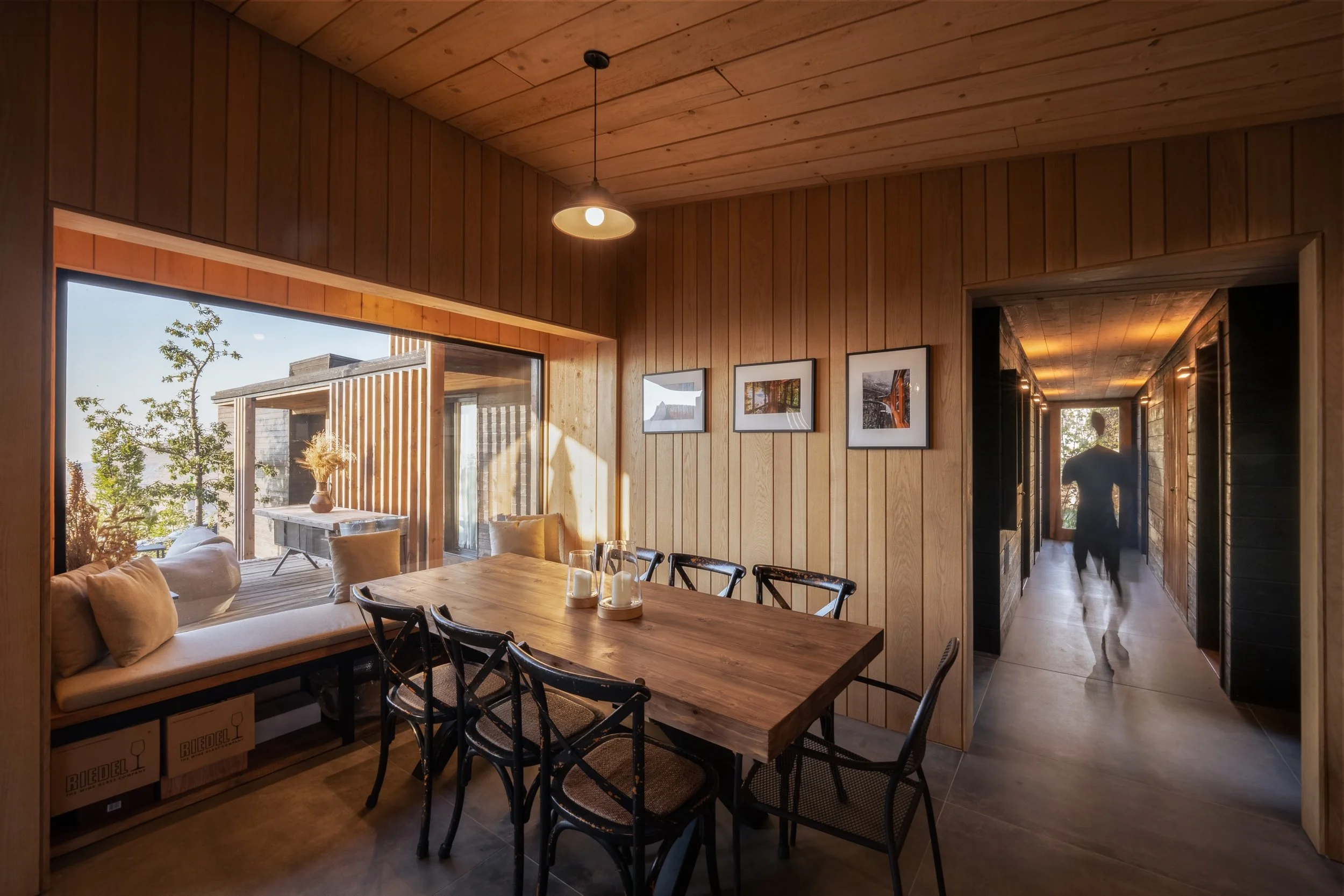 Interior of a wooden house with a dining area. There is a large wooden dining table with multiple chairs, a built-in wooden bench with cushions beside a large window, and framed photos on the wall. A person is visible in the corridor leading outside, and a sliding glass door reveals an outdoor patio with a table and chairs, and a tree.