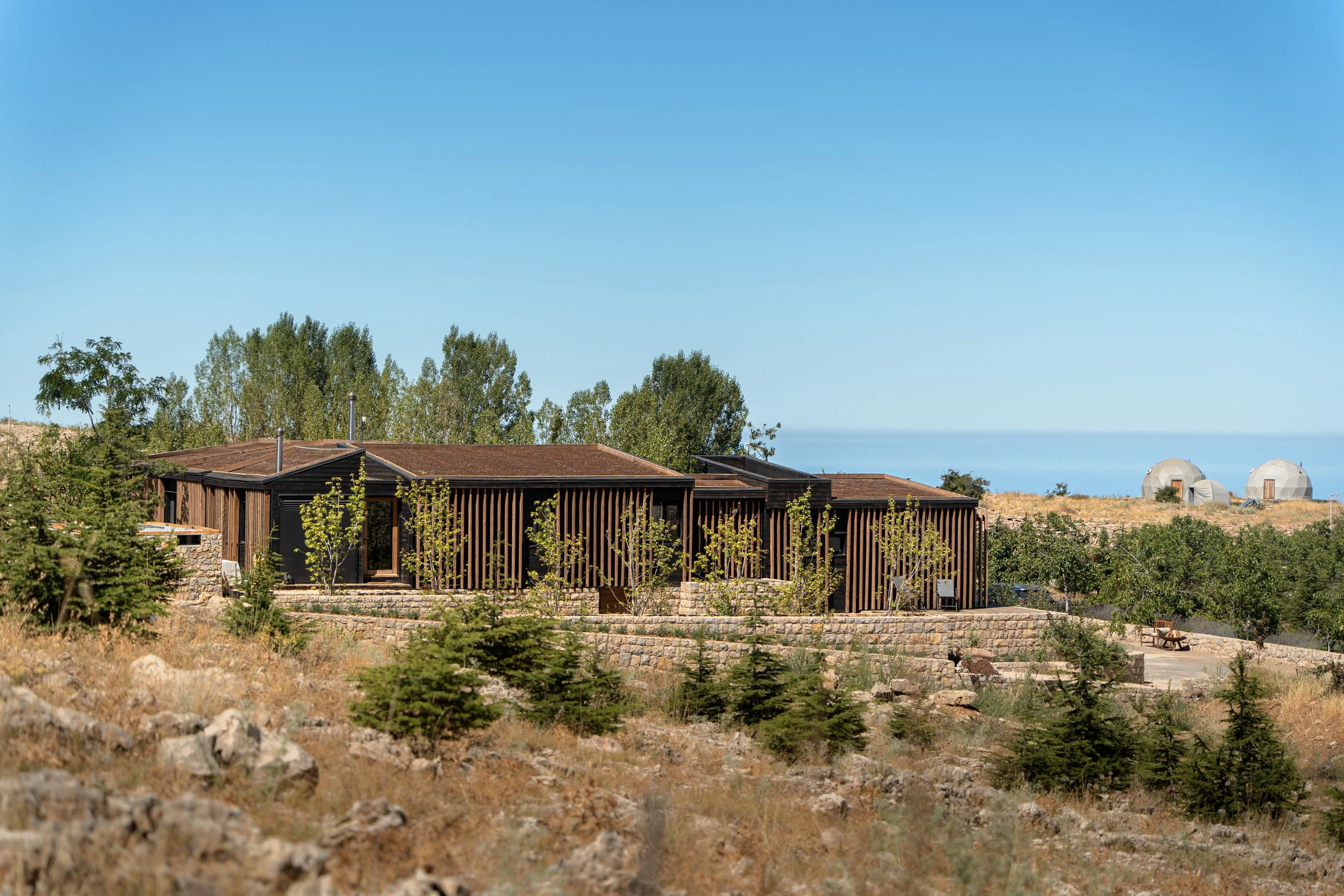 Modern house with wooden slats on the exterior, situated on a dry, rocky landscape with scattered trees, under a clear blue sky.