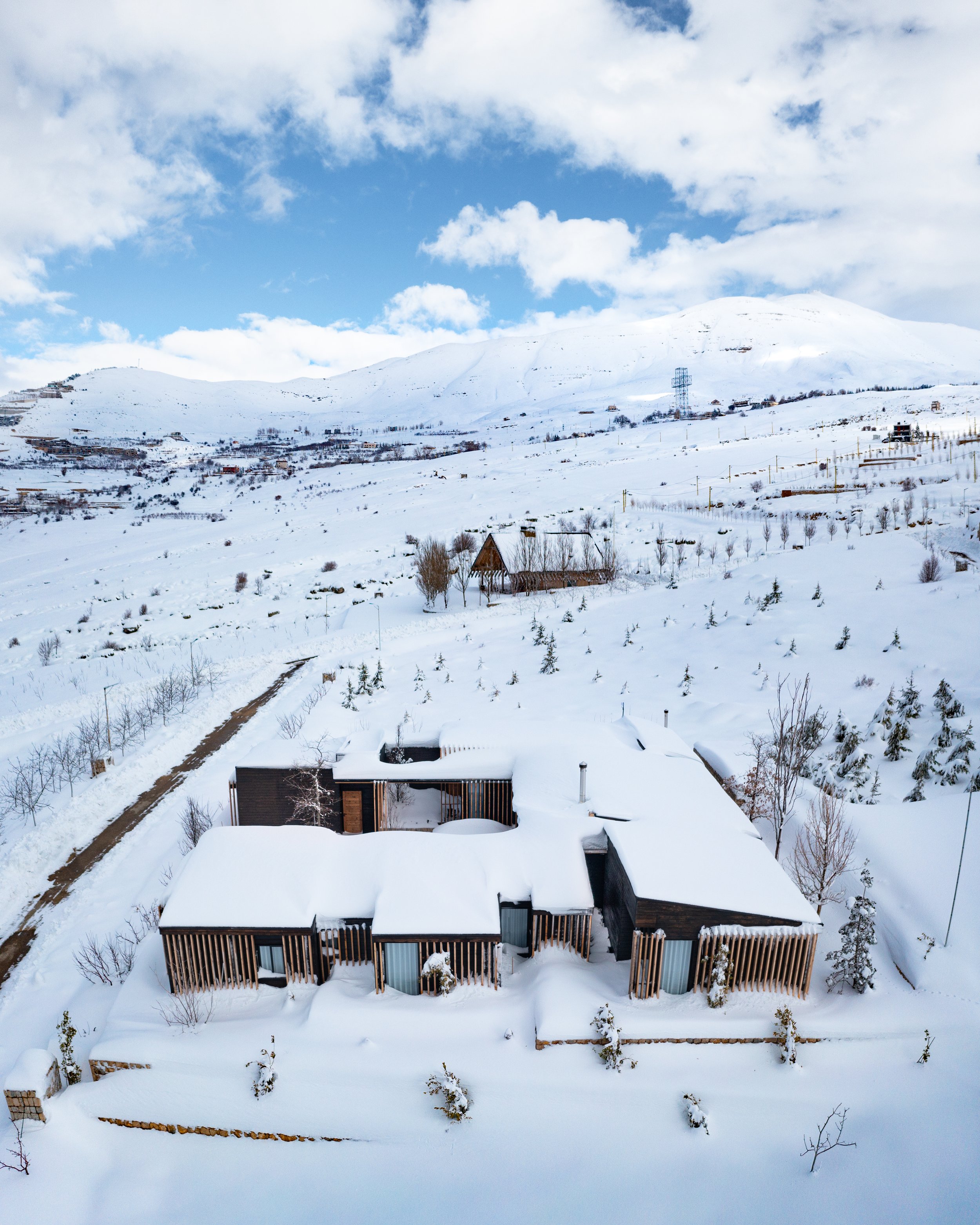 A snow-covered house with a modern design, wooden accents, and large windows, set in a mountainous, snowy landscape with scattered trees and distant hills under a partly cloudy sky.