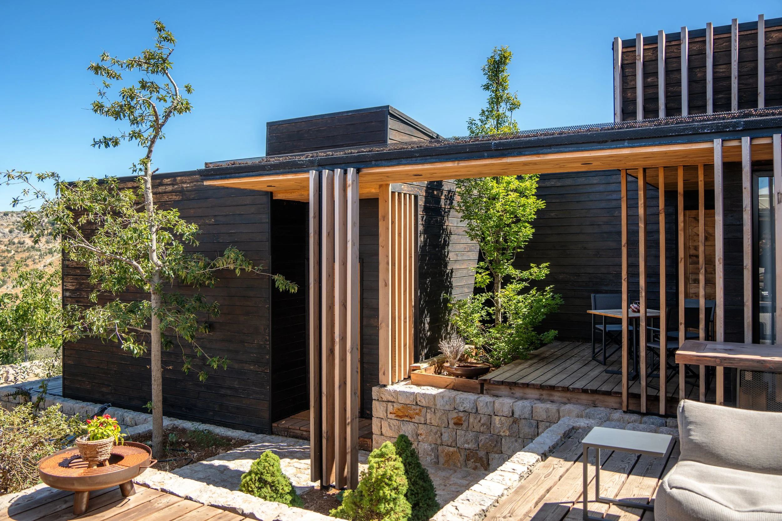 Modern house exterior with wooden and stone elements, small trees, and outdoor seating under a clear blue sky.