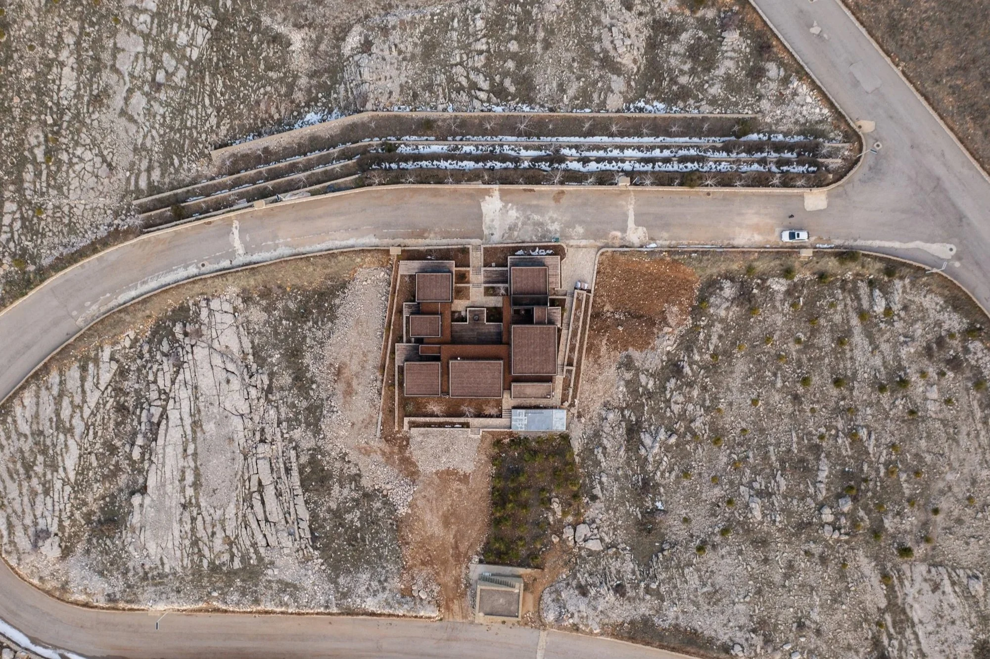 An aerial view of a building surrounded by bare trees and a curved road, with a single white car on the road.