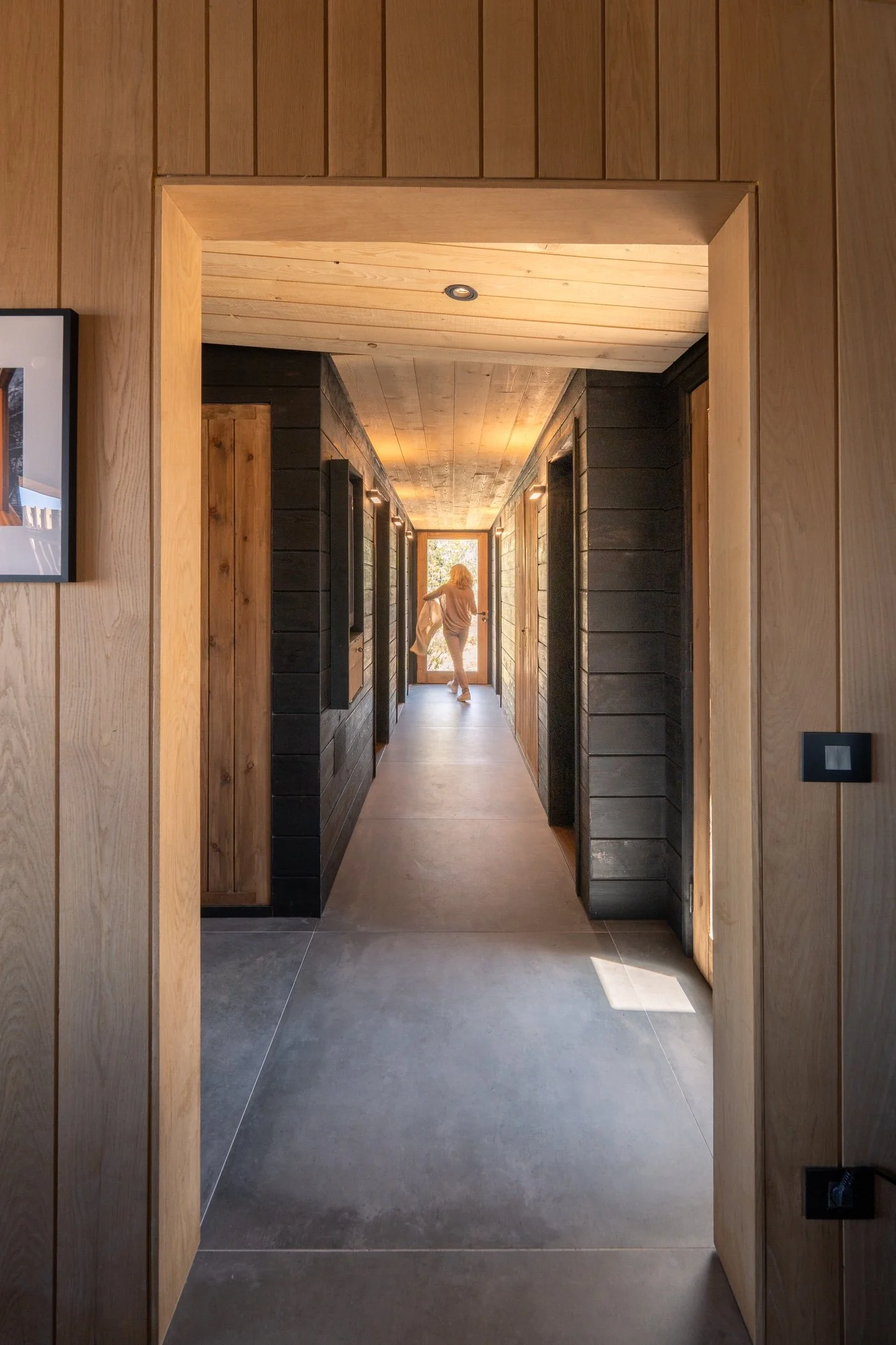 View through a hallway with black wooden walls and natural wood ceiling, lit by recessed lighting, leading to an exit door with sunlight outside.
