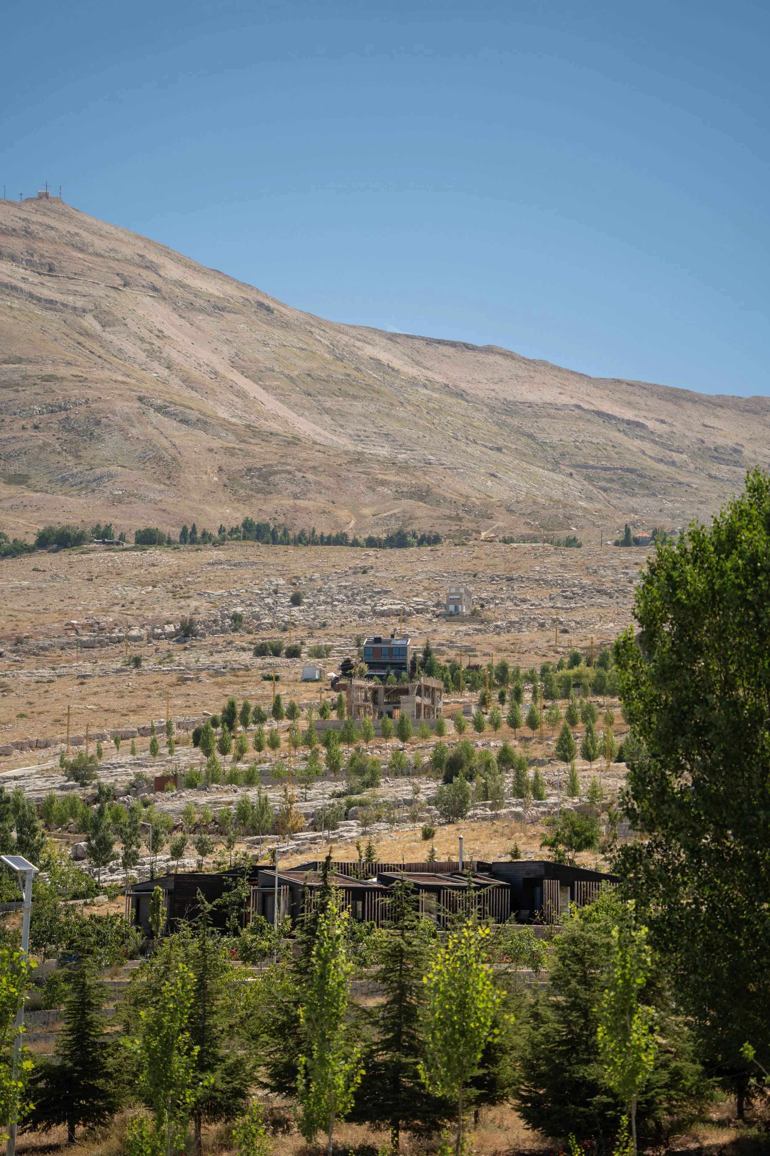 A barren mountain area with sparse vegetation and a few houses, under a clear blue sky.