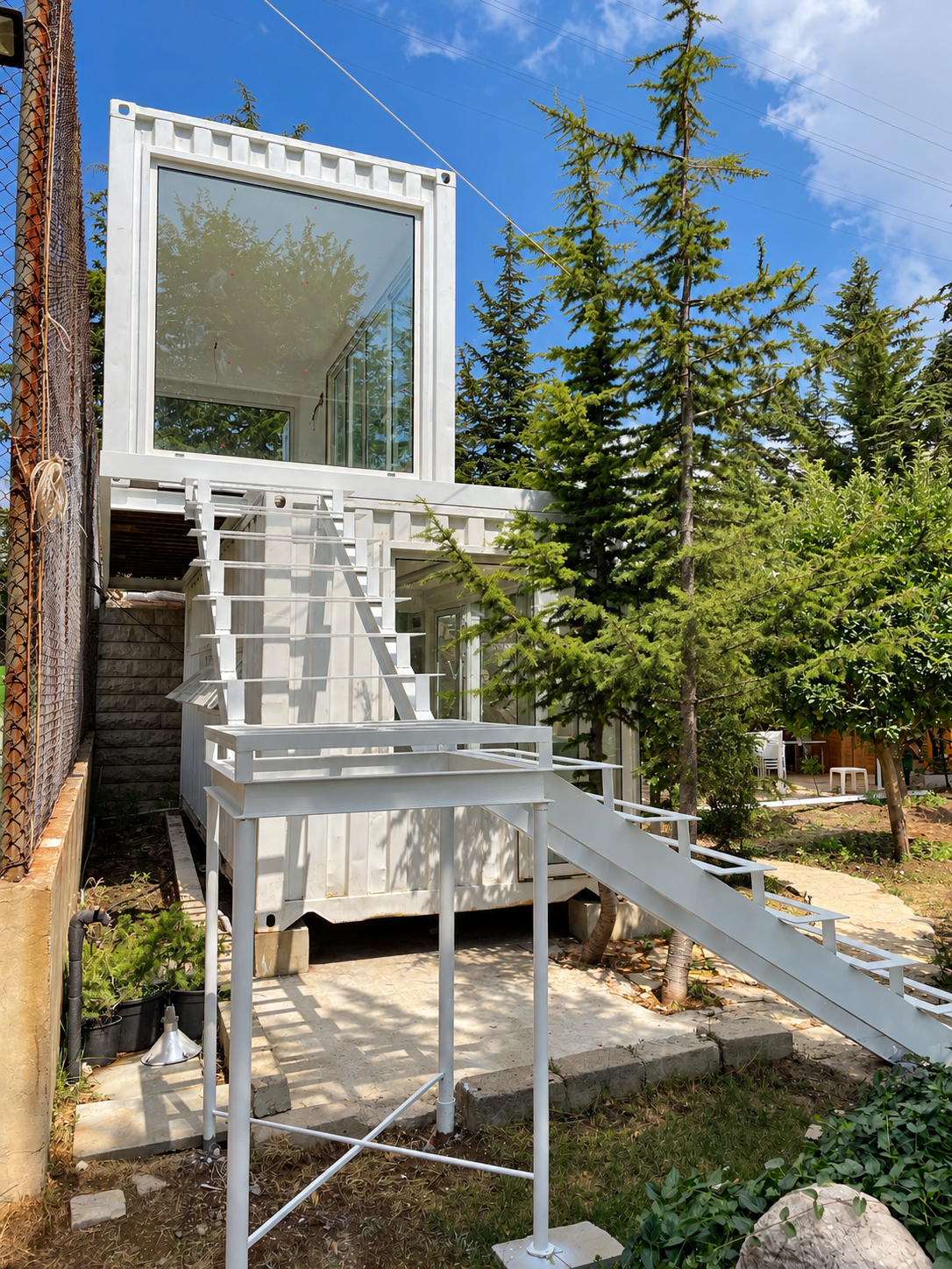 White container building with large glass windows, outdoor staircase with a ramp, surrounded by trees and greenery, under a clear blue sky.