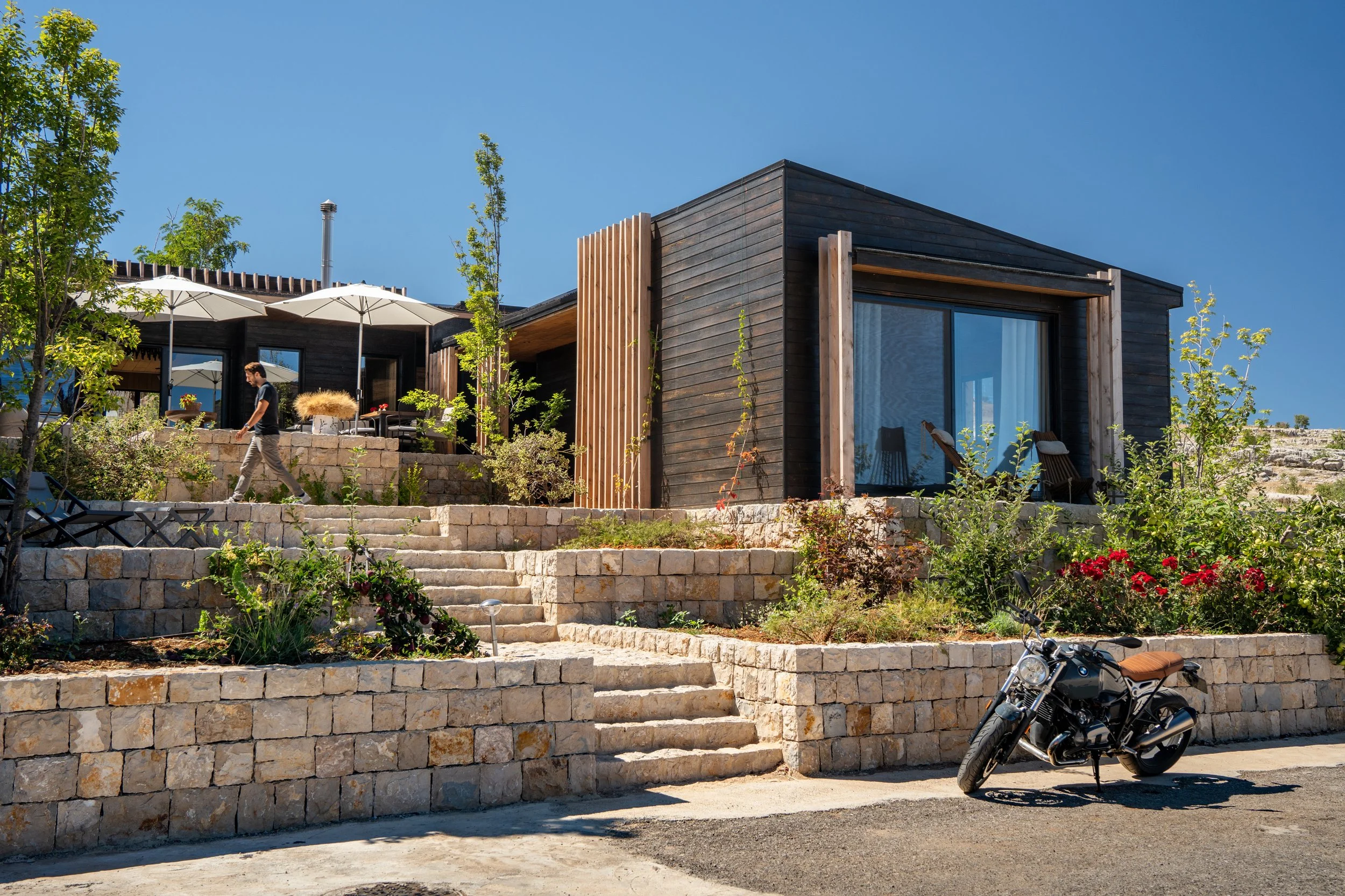 A modern black wooden house with a large sliding glass door, surrounded by plants and trees, with a stone staircase in front. A man is walking on the stairs, and a motorcycle is parked on the ground level.