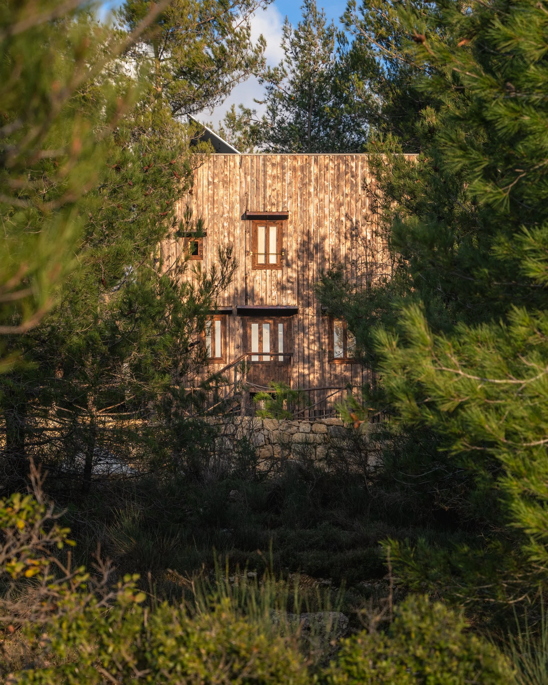 A house with a wooden exterior, seen through green trees and bushes, under a partly cloudy sky.