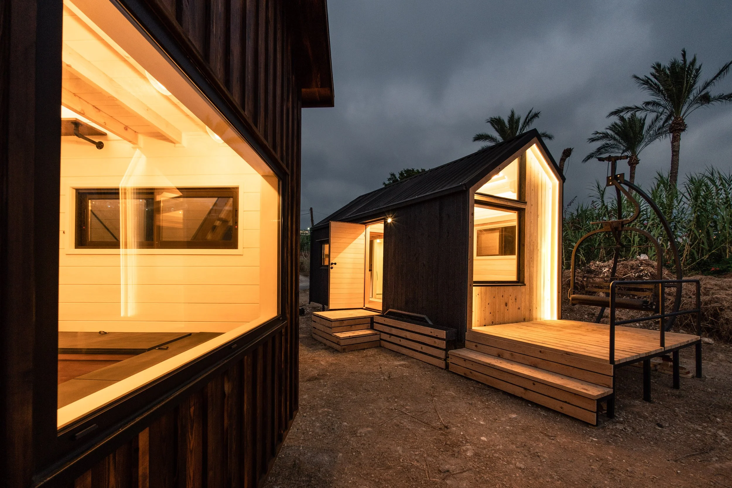 Photo of modern tiny houses with illuminated interiors at dusk, surrounded by palm trees and a cloudy sky.