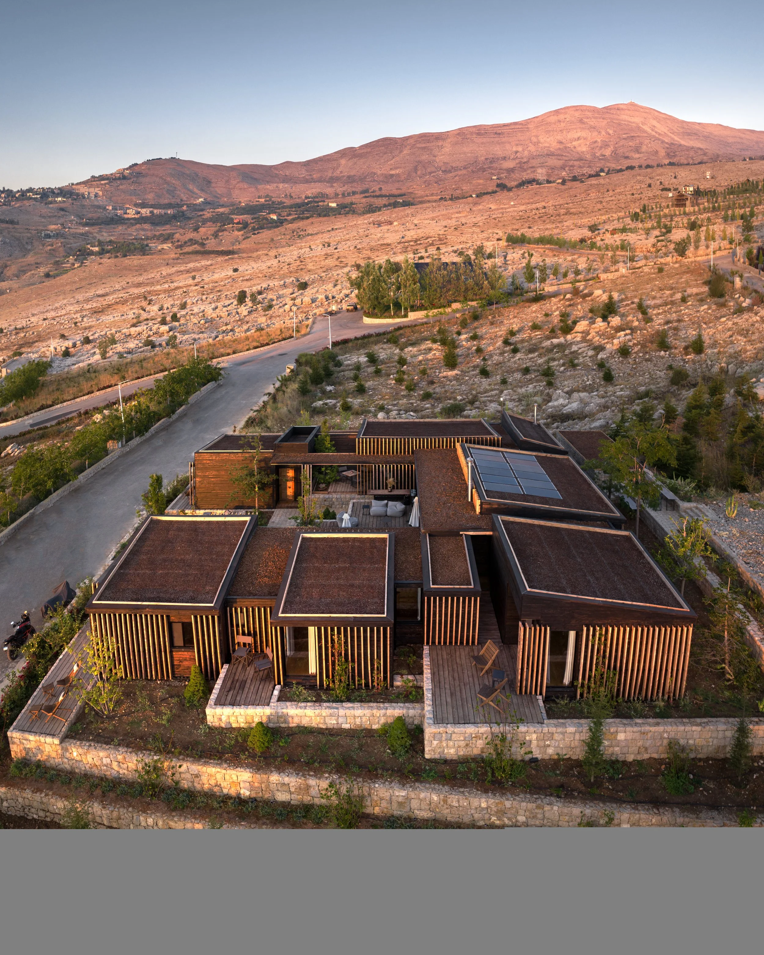Aerial view of modern houses with flat roofs and wooden accents, surrounded by greenery and stone walls, set against a mountainous landscape under a clear sky.