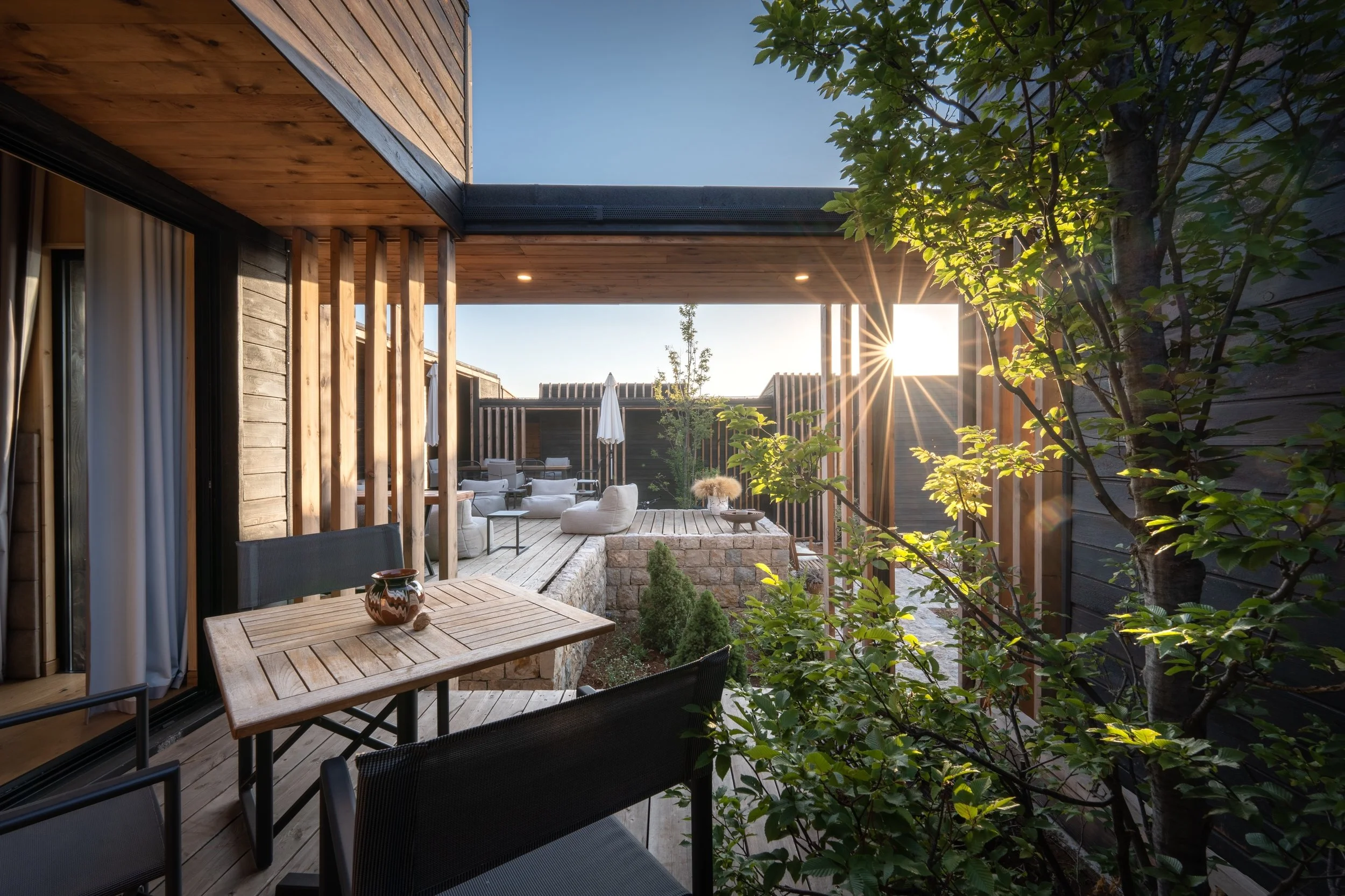 A modern outdoor patio with wooden decking, surrounded by trees and wooden walls, with a seating area including white cushioned chairs, umbrellas, and a wooden table with decorative pottery, bathed in sunlight.